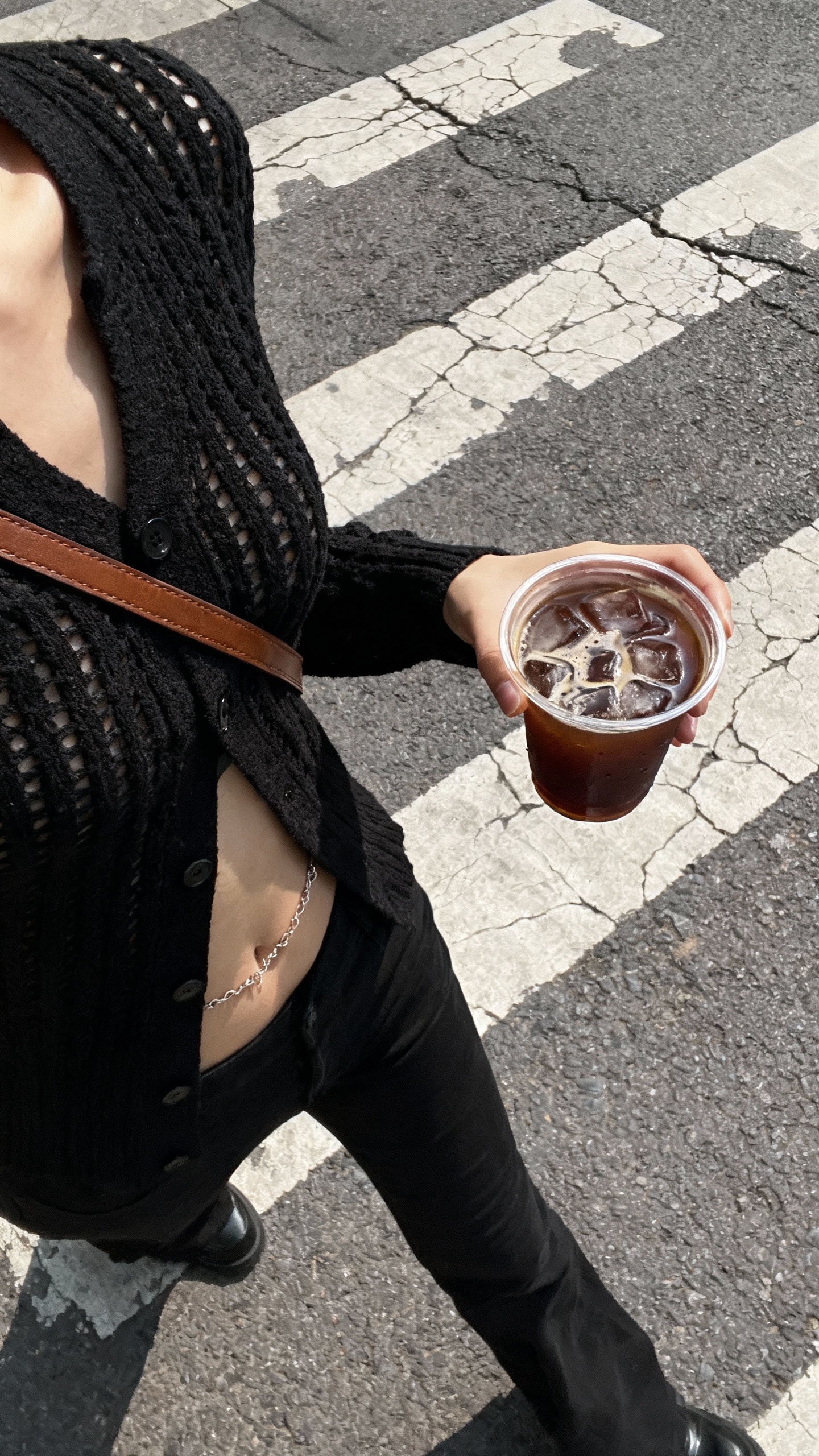 A young, slim woman dressed in a black knit cardigan and a brown crossbody strap is depicted from a direct top-down perspective, capturing her lower torso and right hand prominently in the foreground. The right hand grips a clear cup filled with iced coffee, ice cubes visible through the condensation, while a delicate belly chain peeks subtly from beneath the cardigan. She steps forward on a worn asphalt street marked by cracked white crosswalk stripes, the urban textures sharpening under muted daylight. Shadows cast naturally, emphasizing fabric weaves and the rough surface of the street. The composition conveys a dynamic walking-first-person viewpoint, with focus on tactile textures—the knit of the cardigan, smooth plastic of the cup, and gritty street details—all illuminated softly to evoke an authentic city atmosphere.