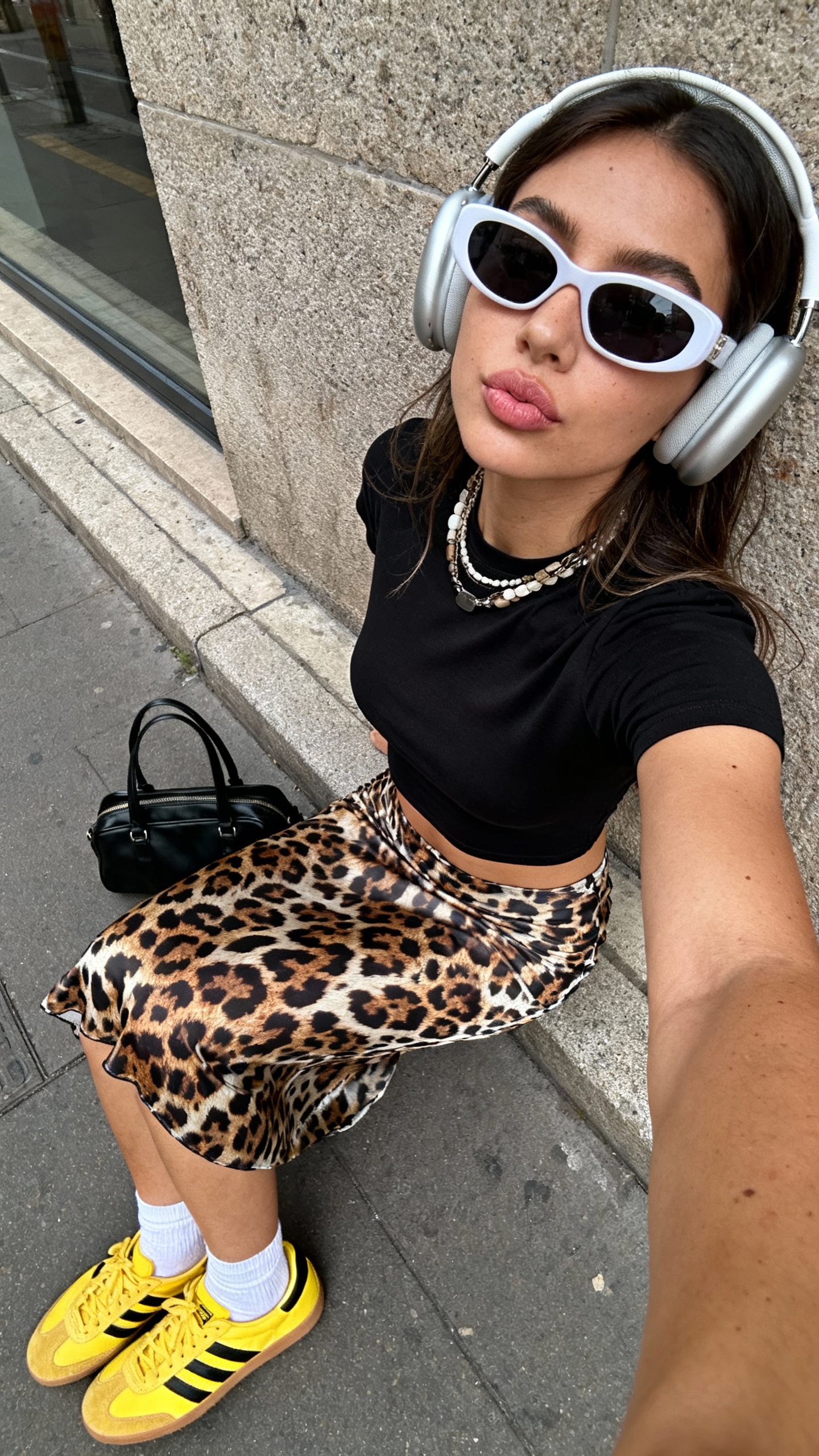 A candid, playfully spontaneous wide-angle iPhone selfie taken from a distinctly elevated overhead angle shows a young woman sitting casually on a city sidewalk ledge, leaning back slightly with her lips softly pursed, directly engaging the camera with a relaxed, neutral expression. She wears an original fitted and cropped black baby tee creatively reimagined without any prints, paired with a uniquely patterned slip skirt inspired by leopard motifs but distinctly stylized with inventive color and texture. Complementing the look are bright yellow sneakers featuring bold black stripes, casual white ankle socks, and an artfully placed black handbag resting on the ground nearby. Her accessories include large, modern headphones, oversized sunglasses with an original shape, and layered necklaces exhibiting varied textures and modern design elements. The authentic urban background features textured stone walls with subtle window reflections and natural daylight casting believable soft shadows and highlights. Textural realism highlights the fabric wrinkles of the tee and skirt, delicate hair strands partially visible under the headphones, natural skin textures with subtle imperfections, and detailed material surfaces of the handbag and sneakers. The composition emphasizes exaggerated wide-angle distortion by enlarging her upper body and face, capturing a spontaneous handheld selfie moment that reflects casual social media aesthetics, self-expression, and stylish urban authenticity.