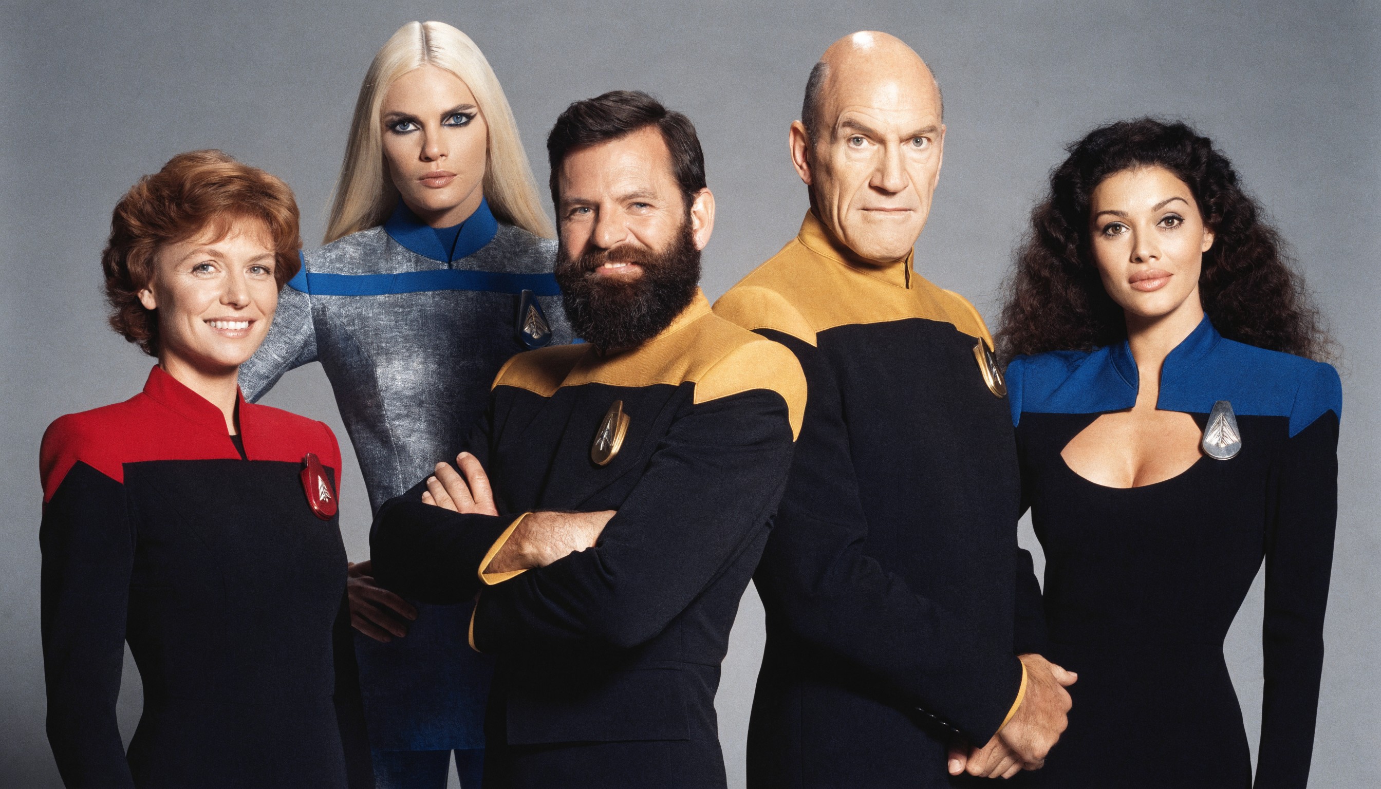 A highly detailed photorealistic studio portrait photograph of five Star Trek: The Next Generation cast members posed closely together against a seamless medium gray backdrop, in the classic 1980s-1990s CBS promotional photo style with soft even studio lighting, subtle shadows, high contrast, and sharp focus on faces. From left to right: 

- First woman (Dr. Beverly Crusher, Gates McFadden likeness): mid-30s Caucasian female with short wavy auburn-reddish brown hair framing her face, warm smile with subtle laugh lines, fair skin, wearing form-fitting black Starfleet uniform with red shoulders and collar, red comm badge on chest, standing confidently with hands at sides.

- Second woman (Lieutenant Tasha Yar, Denise Crosby likeness): late-20s tall athletic blonde Caucasian female with long straight platinum blonde hair parted in center falling past shoulders, striking dramatic black eye makeup accentuating her blue eyes, neutral confident expression, pale skin, wearing sleek gray-silver toned Starfleet sciences uniform with blue shoulders, blue comm badge, posed slightly behind with one hand on hip.

- Central man (Commander William Riker, Jonathan Frakes likeness): early-40s tall broad-shouldered Caucasian male with short dark brown hair, full thick dark beard and mustache, intense smiling expression with crinkled eyes, tanned skin, wearing black Starfleet uniform with mustard-yellow/gold shoulders and collar, gold comm badge, arms crossed confidently.

- Next man (Captain Jean-Luc Picard, Patrick Stewart likeness): mid-50s bald Caucasian male with shiny smooth bald head, prominent ears, stern yet warm smiling expression with thin lips and high cheekbones, light skin, wearing black Starfleet uniform with mustard-yellow/gold shoulders and collar, gold comm badge, standing with hands clasped in front.

- Rightmost woman (Counselor Deanna Troi, Marina Sirtis likeness): late-20s curvaceous Mediterranean female with long voluminous dark brown nearly black curly hair cascading over shoulders, large expressive dark eyes, full lips in soft smile, olive skin, wearing elegant black Starfleet uniform dress with deep blue shoulders, low scoop neckline revealing subtle cleavage, blue comm badge, posed gracefully with hands relaxed at sides.

All actors in precise iconic 1987 Season 1 poses, slight overlapping arrangement in a tight group formation, neutral professional expressions with subtle camaraderie, rich color saturation on uniforms (crisp black fabrics, vibrant red/blue/gold panels with subtle sheen), natural skin tones, high-resolution 35mm film grain texture, perfect anatomical accuracy, dramatic yet approachable composition, 8k ultra-detailed, cinematic depth of field with razor-sharp faces.