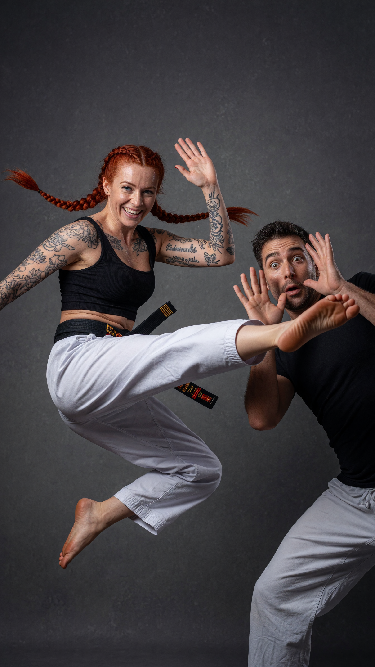A highly detailed realistic studio photograph of a dynamic martial arts action scene against a seamless dark gray mottled backdrop: a smiling athletic woman with vibrant red hair in two long braids, blue eyes, and extensive black floral and script tattoos covering both arms, wearing a black ribbed crop top, white karate gi pants with a black belt featuring gold and red stripes, and barefoot, captured mid-air executing a powerful high side kick with her right leg fully extended toward the man's face; a surprised man with short dark hair, stubble, and wide eyes wearing a black fitted t-shirt and matching white karate pants, defensively raising both hands to block or touch her incoming foot while leaning back slightly; dramatic studio lighting with soft shadows, sharp focus, photorealistic textures on skin, fabric folds, and hair strands, cinematic composition, high resolution, professional martial arts portrait style.