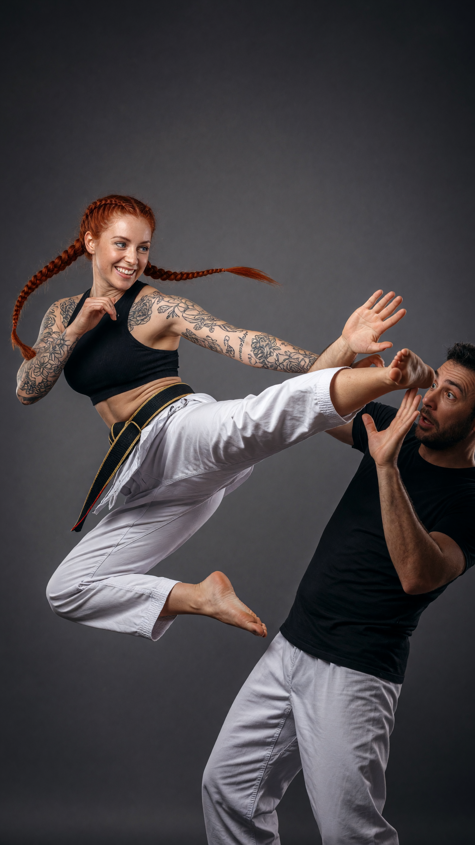 A highly detailed realistic studio photograph of a dynamic martial arts action scene against a seamless dark gray mottled backdrop: a smiling athletic woman with vibrant red hair in two long braids, blue eyes, and extensive black floral and script tattoos covering both arms, wearing a black ribbed crop top, white karate gi pants with a black belt featuring gold and red stripes, and barefoot, captured mid-air executing a powerful high side kick with her right leg fully extended toward the man's face; a surprised man with short dark hair, stubble, and wide eyes wearing a black fitted t-shirt and matching white karate pants, defensively raising both hands to block or touch her incoming foot while leaning back slightly; dramatic studio lighting with soft shadows, sharp focus, photorealistic textures on skin, fabric folds, and hair strands, cinematic composition, high resolution, professional martial arts portrait style.