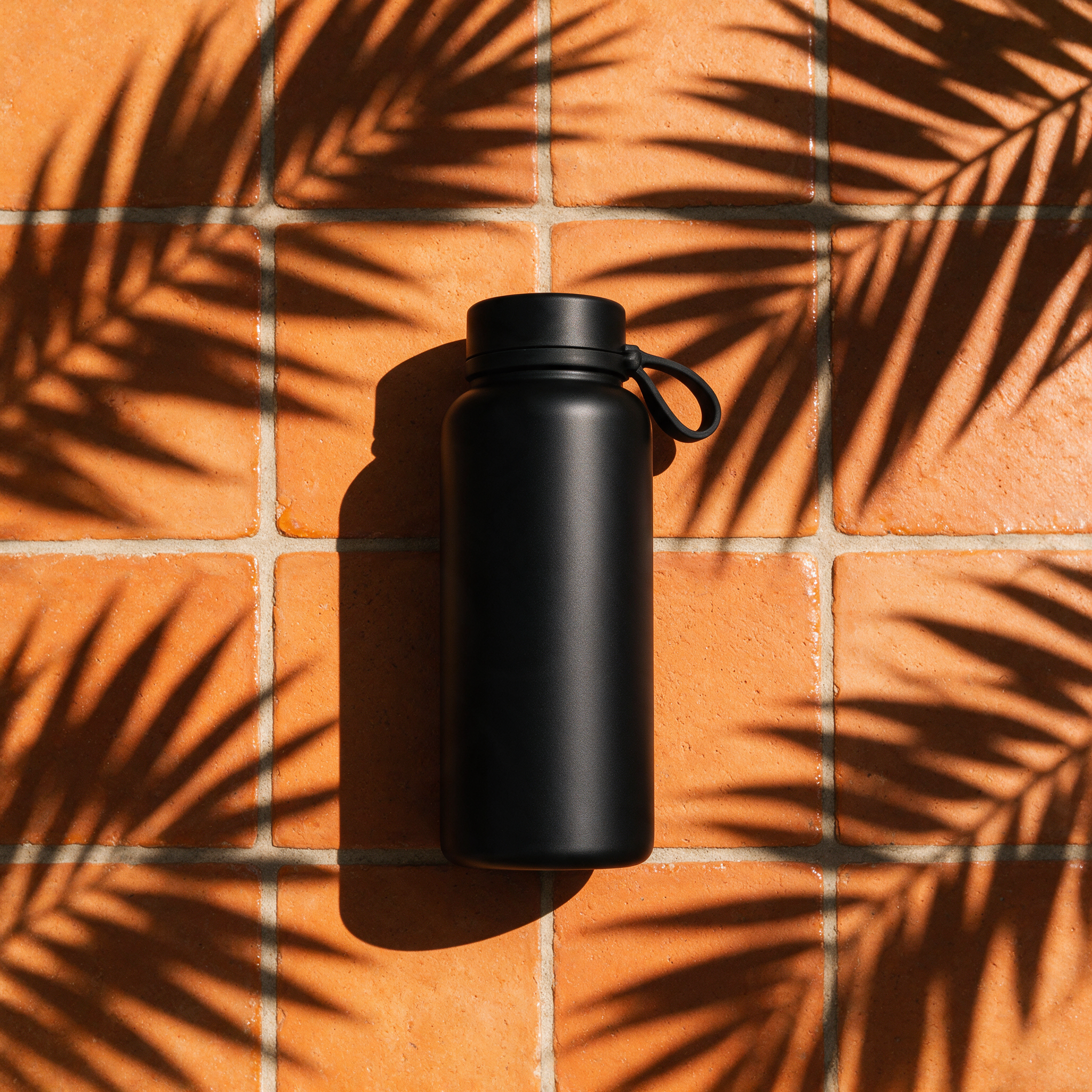 a flat-lay product hero shot of a matte black water bottle on terracotta tiles with sharp palm-leaf shadows, studio lighting, ad-ready composition