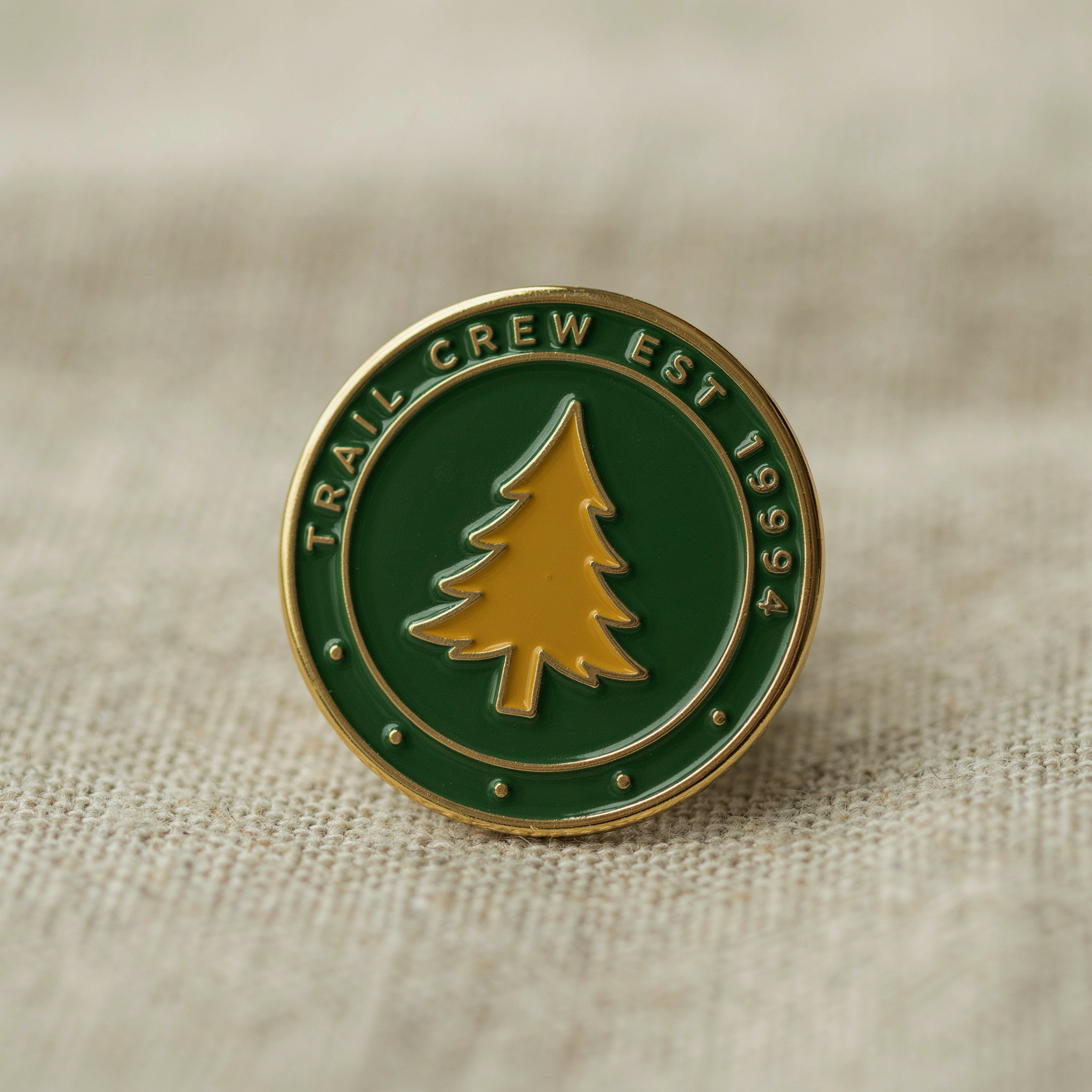 An enamel pin design laid on linen, circular badge reading "TRAIL CREW EST 1994" around a pine tree emblem, forest green and mustard, soft studio light