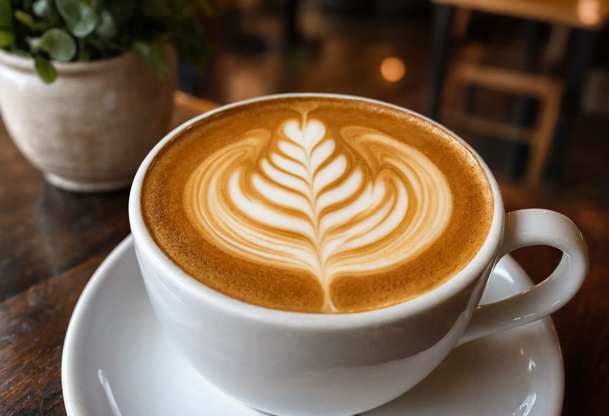 A close-up of an artisan latte with intricate rosetta foam art in a white cup, warm cafe background blurred, inviting