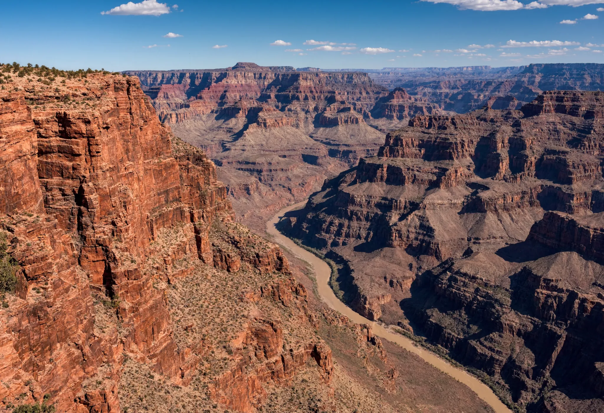 A sweeping desert canyon at midday, layered red rock formations, deep shadows, a narrow river far below, epic landscape scale