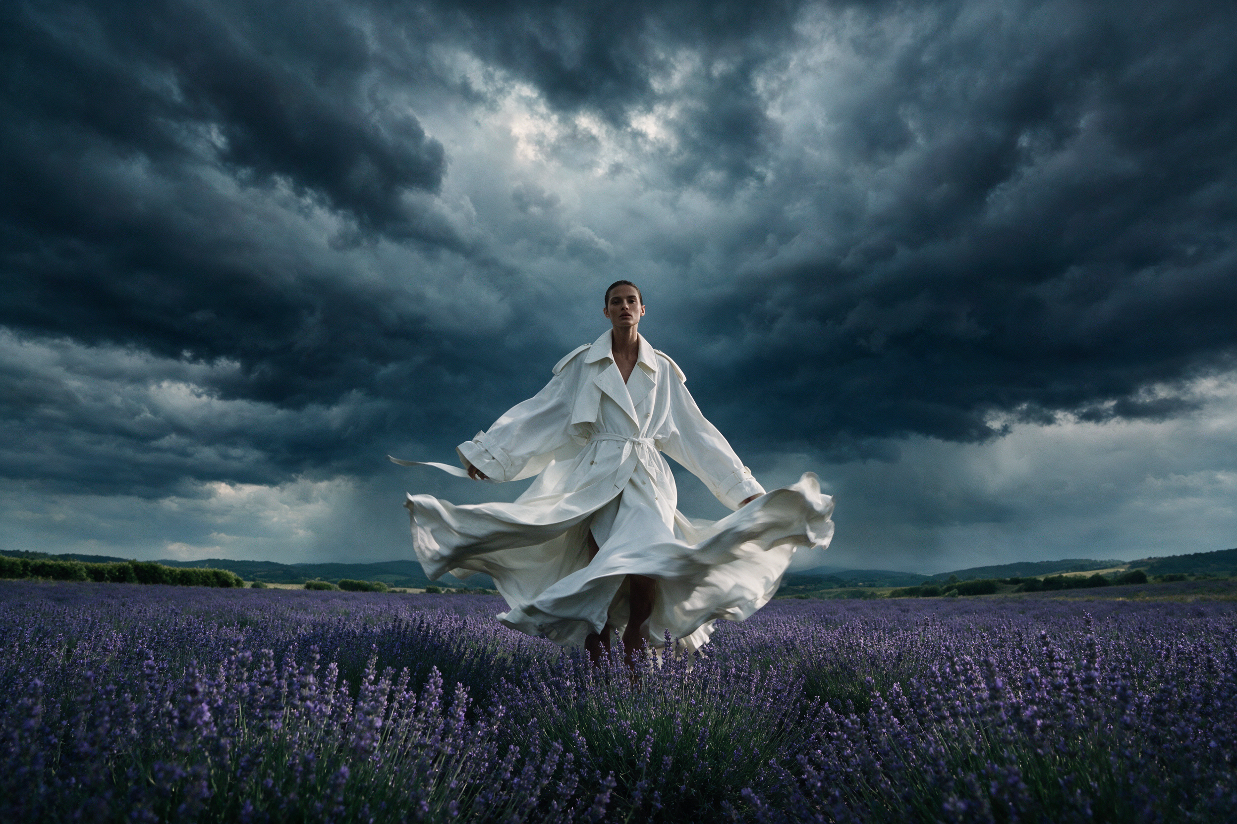 Fashion editorial of a model in an oversized white trench coat standing in a field of lavender under a stormy sky, wind-blown fabric, high-fashion magazine look