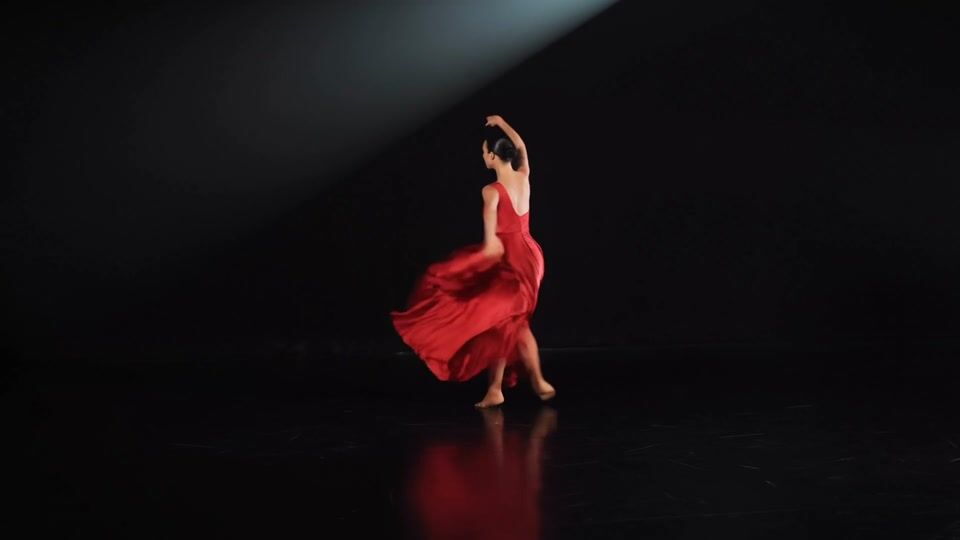 A dancer with a sleek black bun spins in a flowing crimson silk gown on a black stage, the skirt flaring into a wide red arc under a single white spotlight
