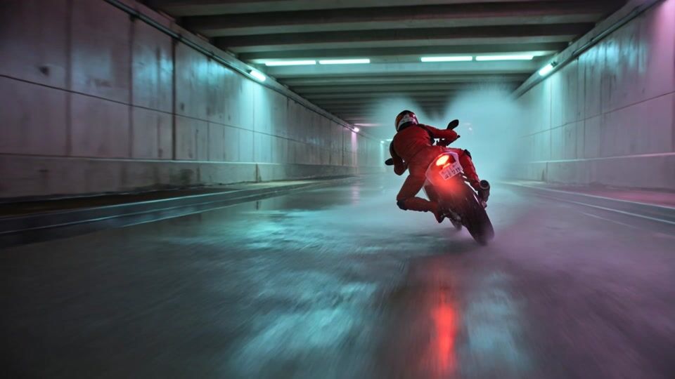 A rider in a red leather suit leans a motorcycle through a flooded concrete underpass at night, a tall fan of spray kicking off the rear tire under cyan and magenta tube lights as she speeds down the tunnel