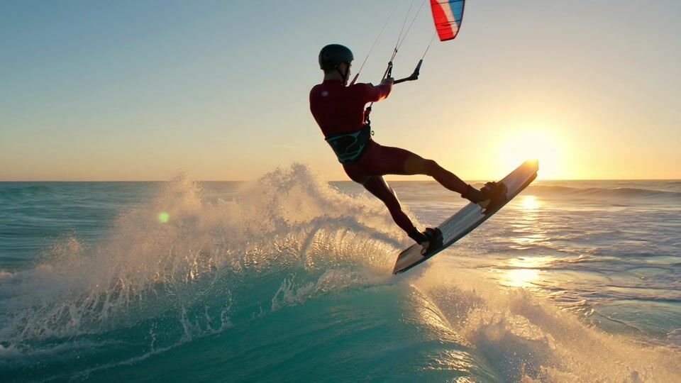 A kitesurfer in a red wetsuit carves up a turquoise wave at sunset and launches into the air, spray lit gold by the low sun behind him