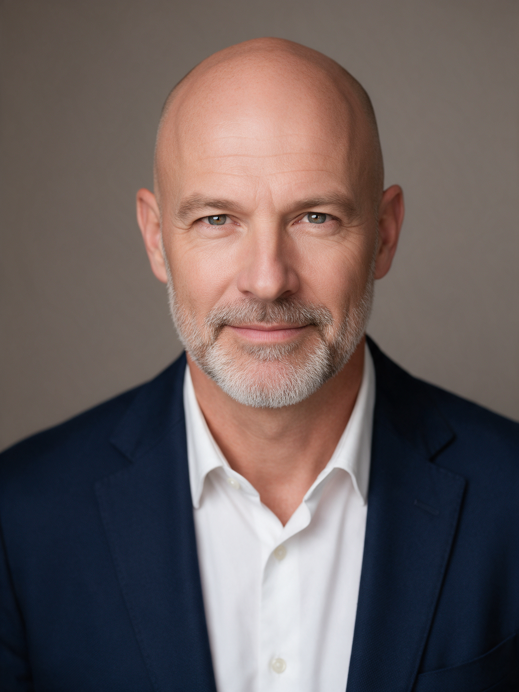 A bald man with a trimmed grey beard in a navy blazer and white shirt against a warm grey backdrop