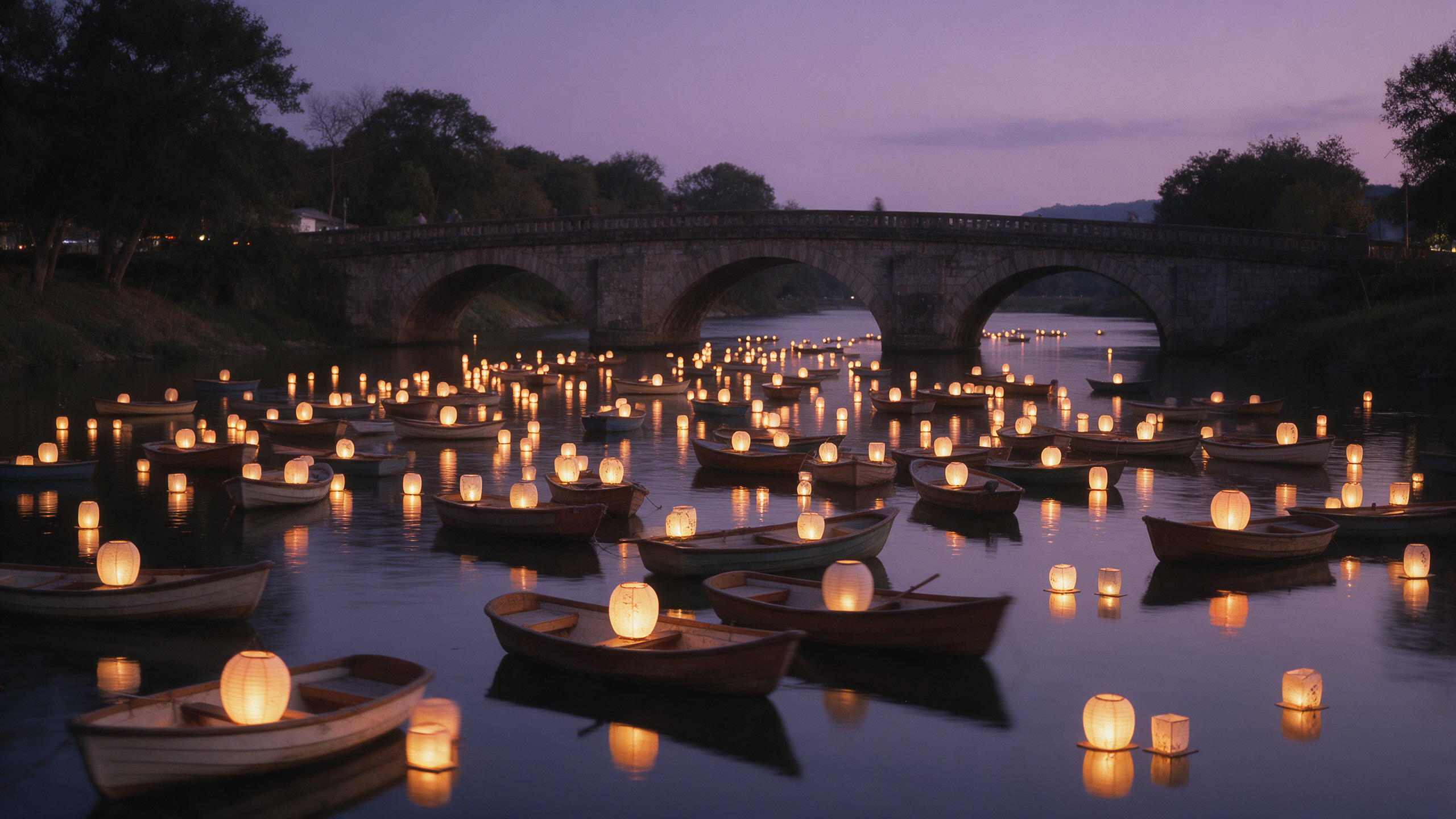 Dozens of small rowboats with glowing lanterns drift on a calm river at dusk below a stone arch bridge