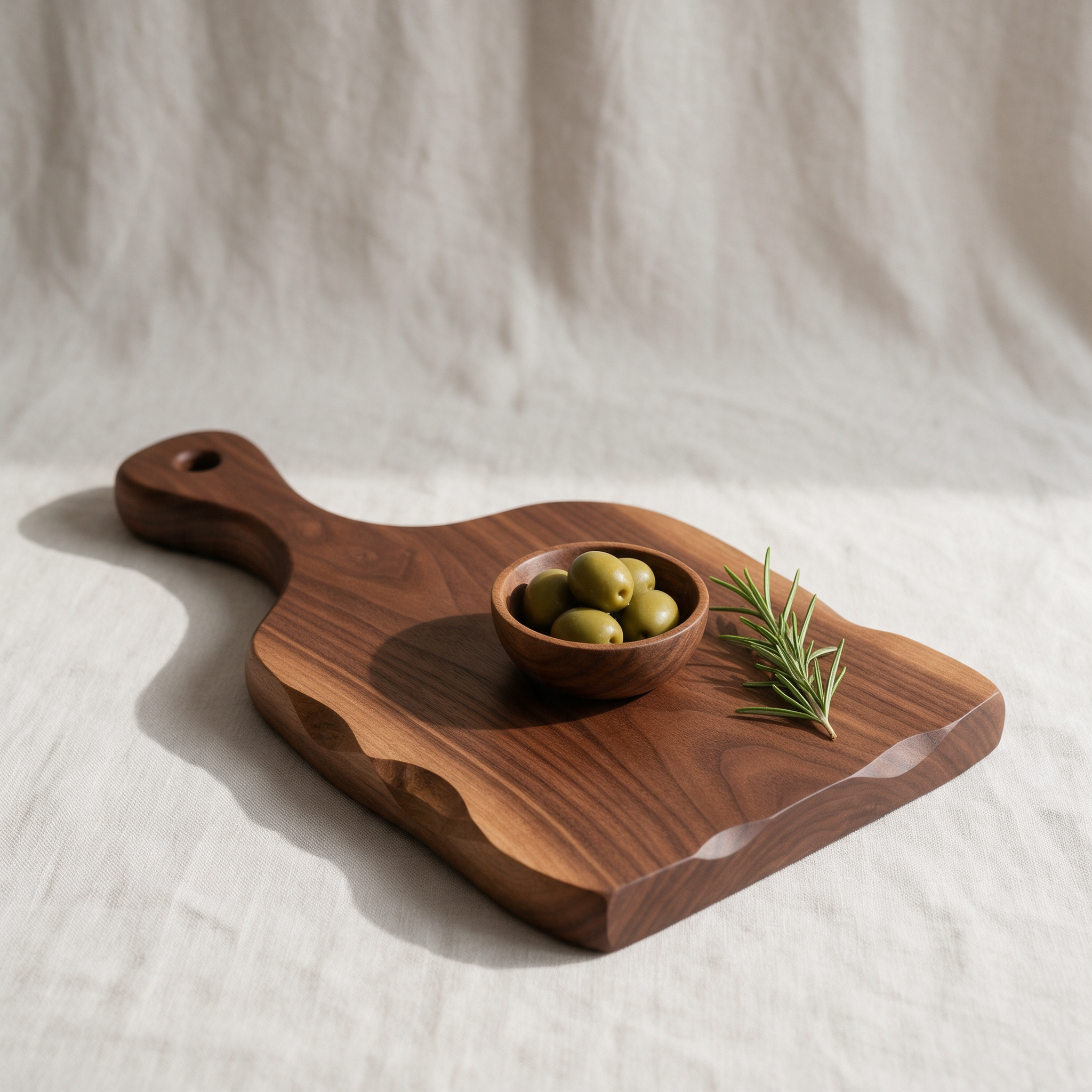 A carved walnut serving board with green olives and rosemary on pale linen in soft window light