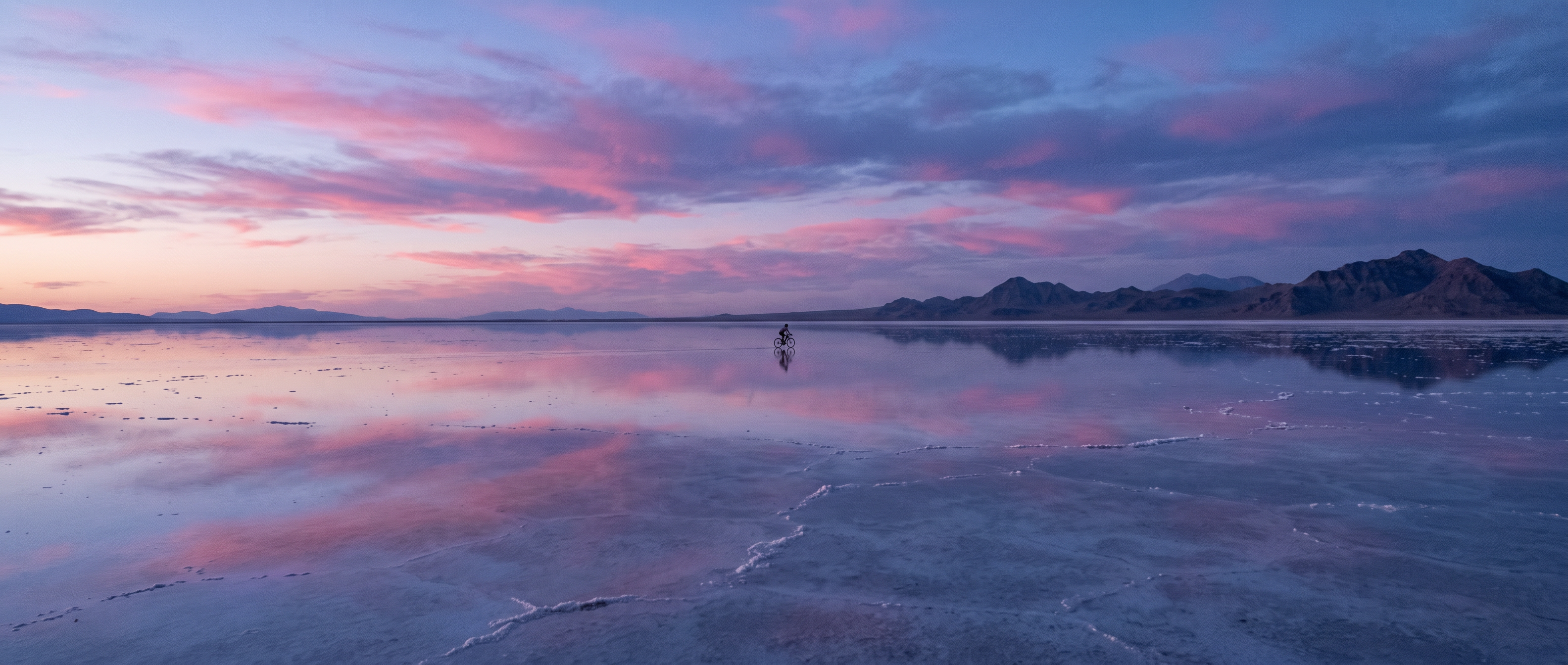 An ultra wide salt flat at dusk mirroring pink and violet clouds with a tiny cyclist on the horizon