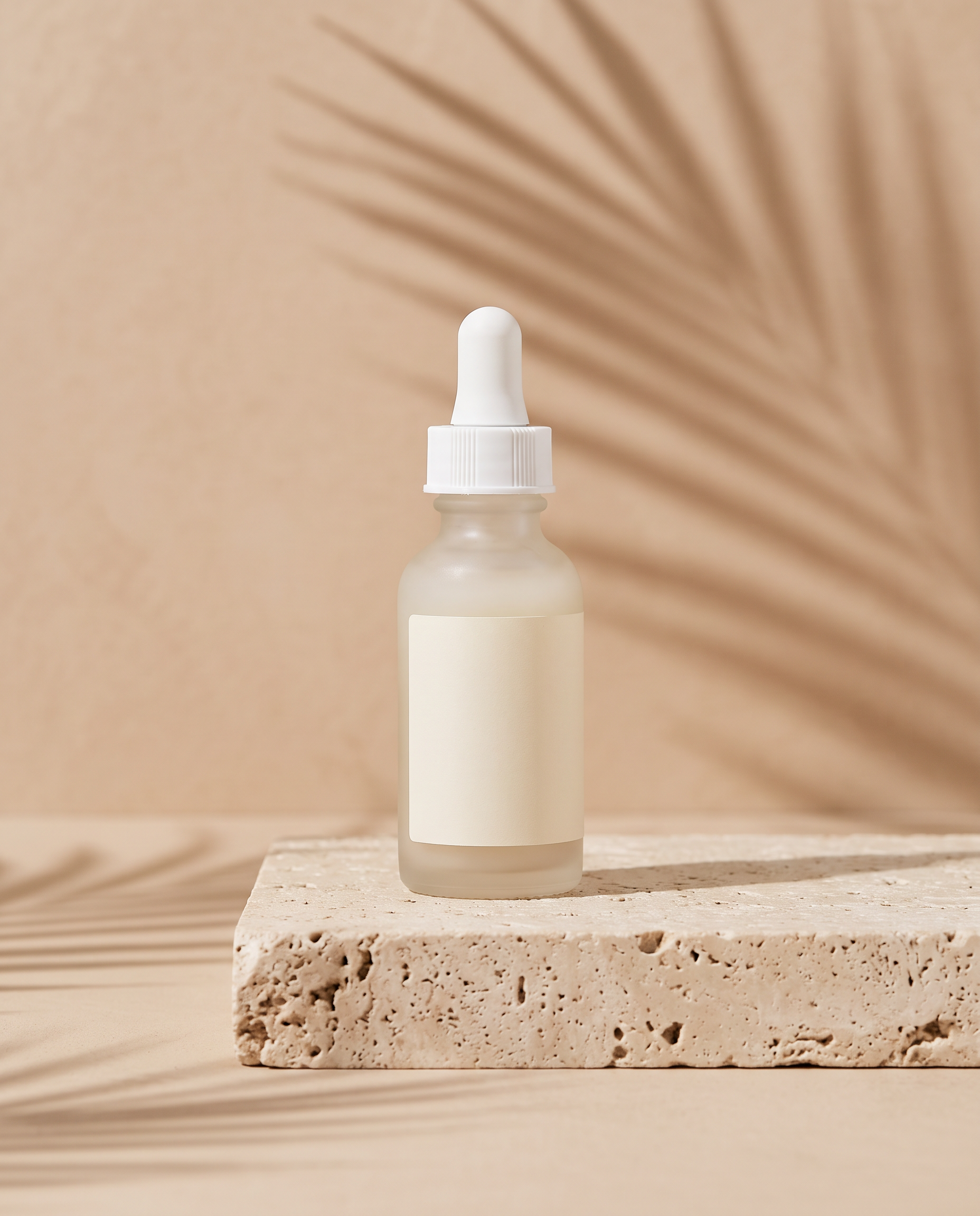 A frosted serum bottle with a blank label on a travertine plinth with palm shadows