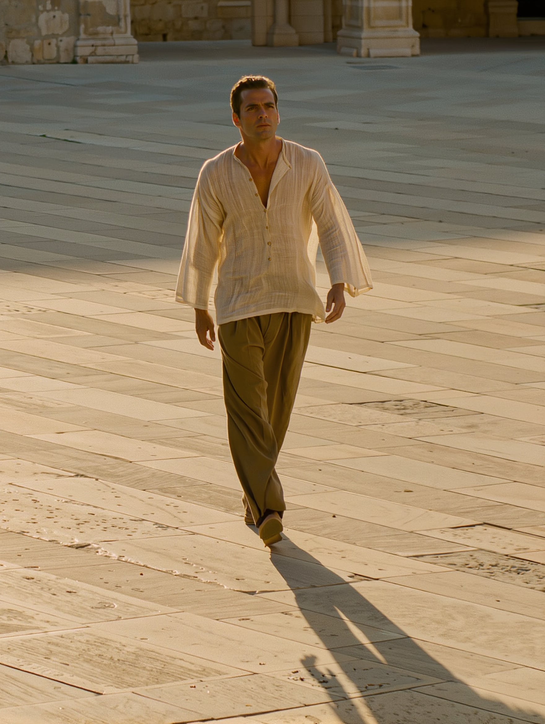 A man in a cream linen shirt and olive trousers walks across a sunlit stone plaza with a long shadow