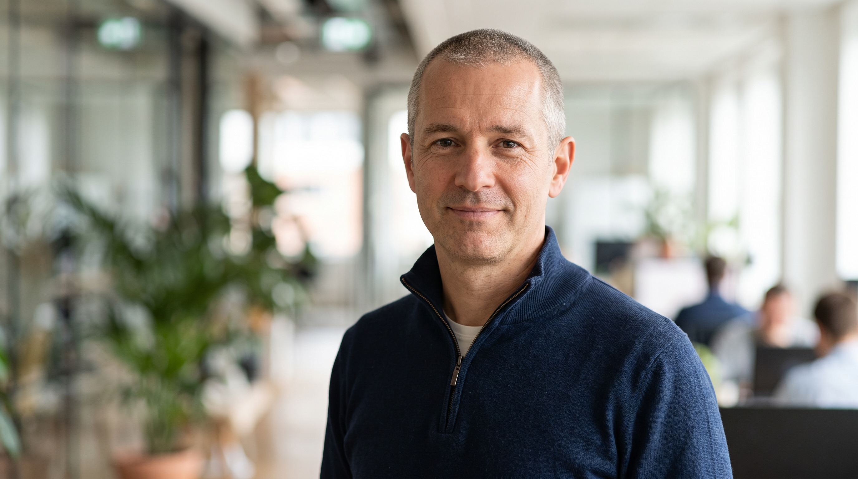 A trainer in his forties with close cropped grey hair and a navy quarter zip smiles at the camera in a bright office