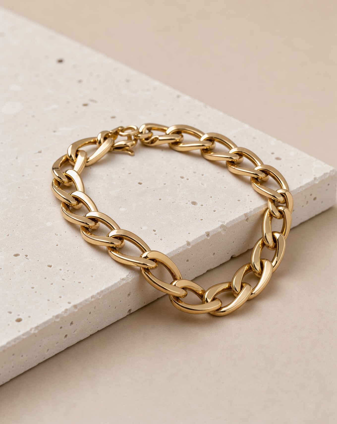 A chunky gold chain bracelet draped over white travertine with crisp reflections on each link
