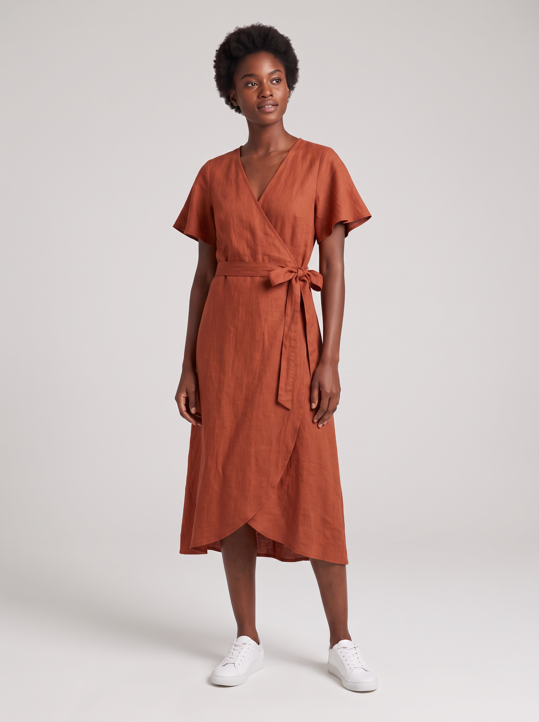 A model with short natural curls stands on a grey studio backdrop in a rust linen wrap midi dress with flutter sleeves, a tie waist and white sneakers
