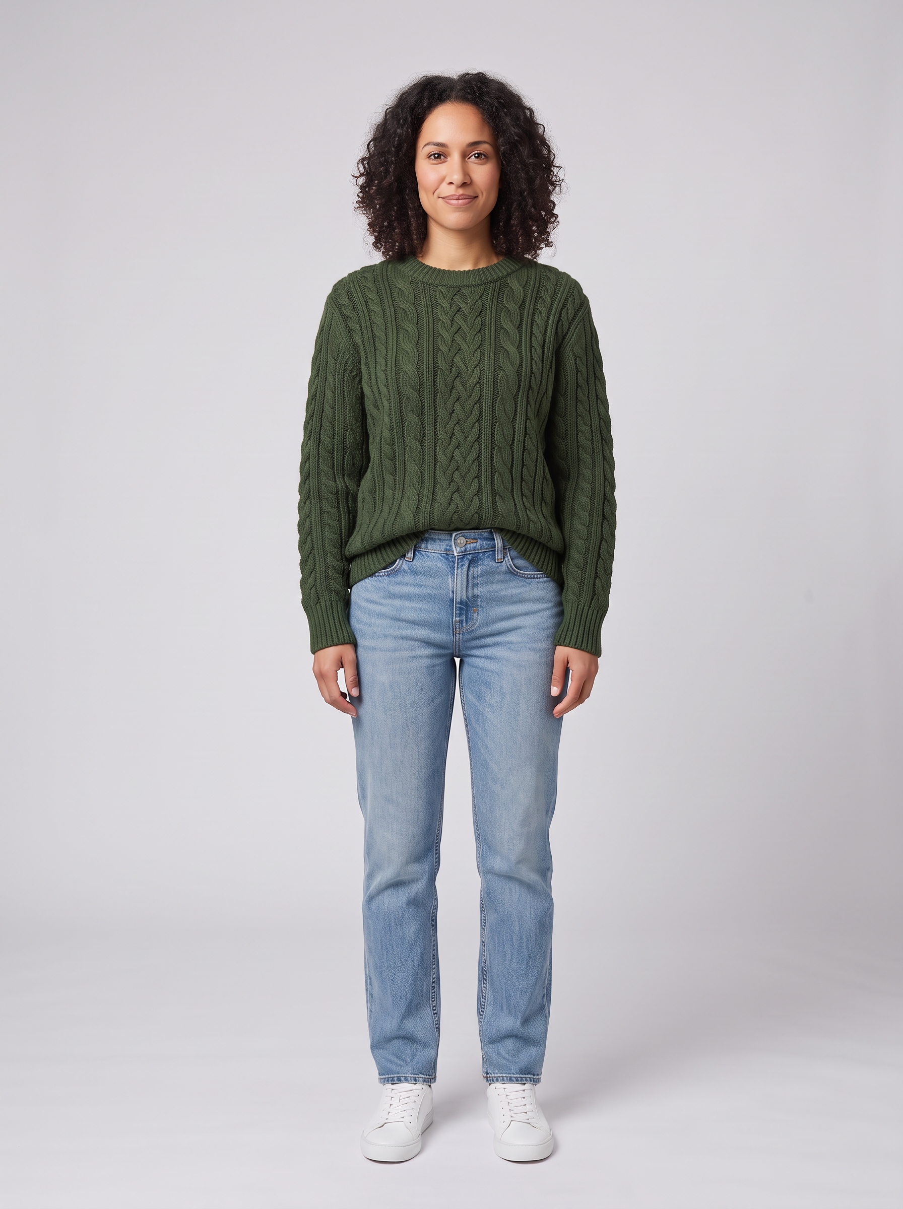 The same woman in a forest green cable knit sweater over her jeans and white sneakers on grey