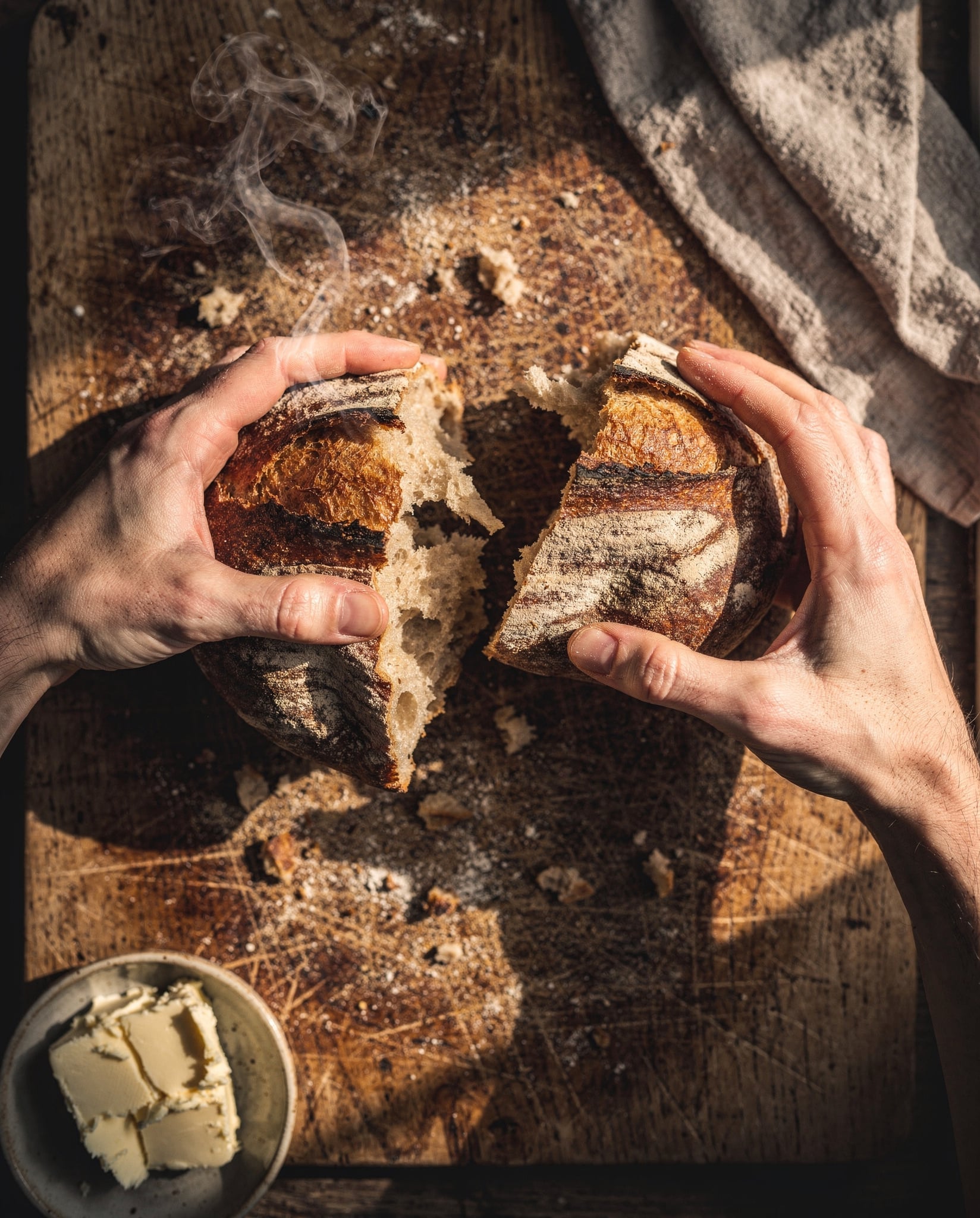 Overhead view of two flour dusted hands tearing a dark crusted sourdough loaf apart on a scarred oak board, steam curling from the open crumb, crumbs scattered, a dish of butter in the corner and a linen towel at the top edge in low side light