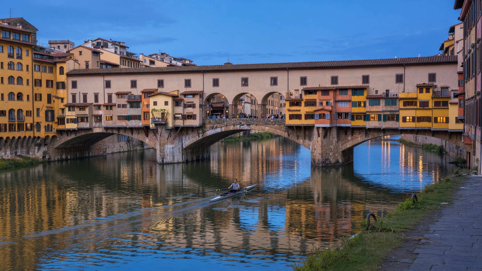 The Ponte Vecchio in Florence at blue hour, the old shops along the bridge lit warm above the still Arno river and its reflections, a single rowing scull gliding toward the arches