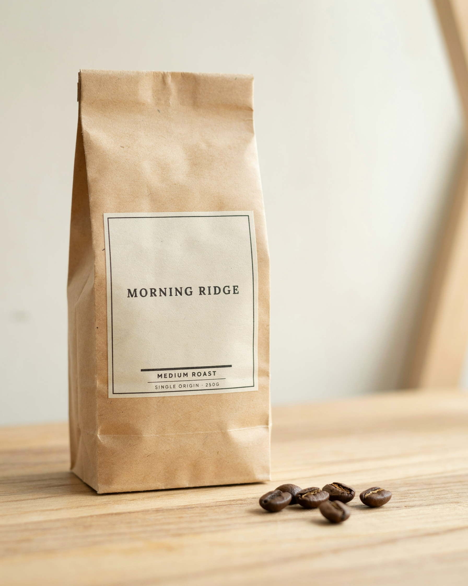 A kraft paper coffee bag with a cream label reading MORNING RIDGE, MEDIUM ROAST and SINGLE ORIGIN 250G on an oak shelf beside a few beans