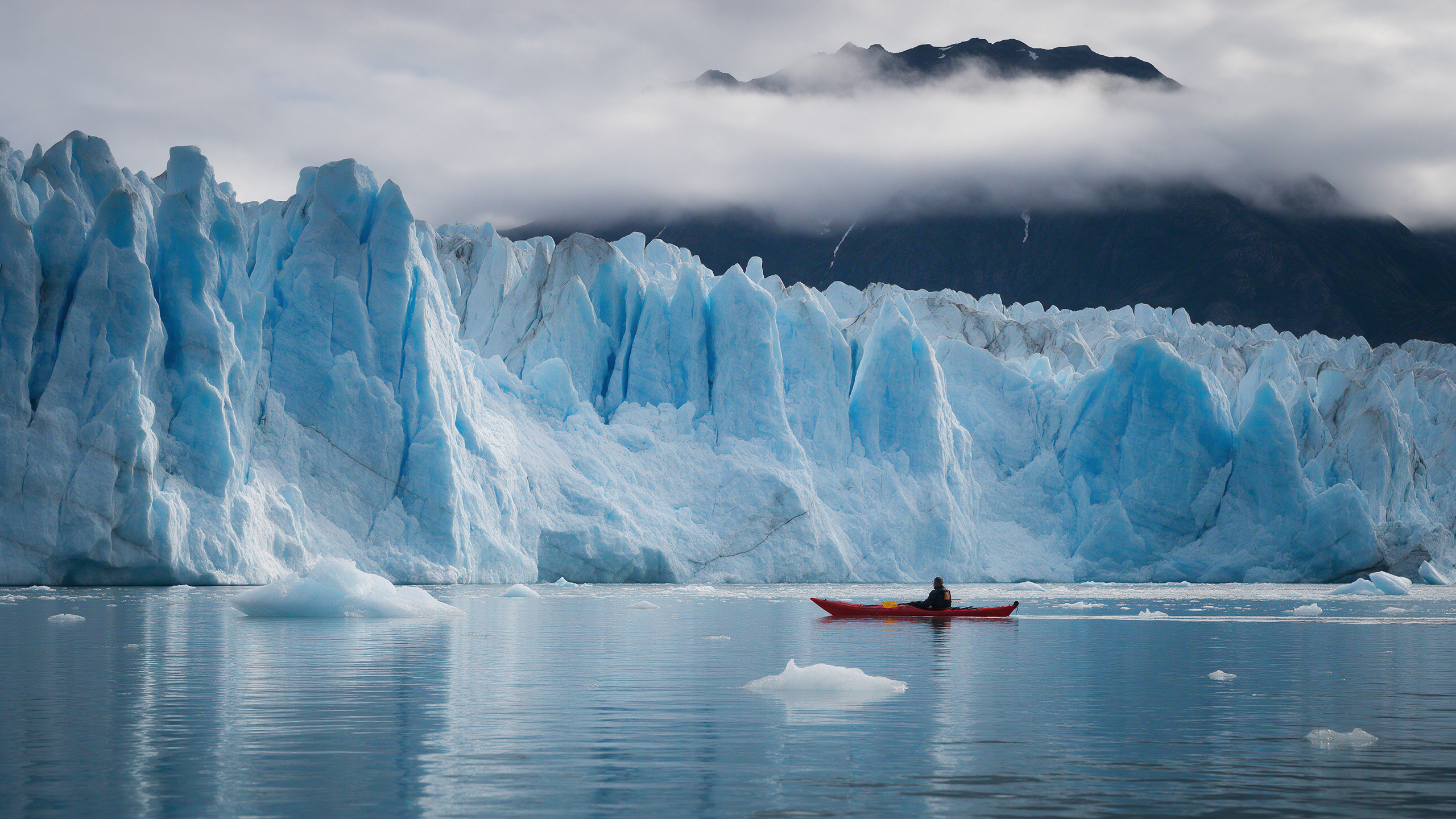 A lone red kayak glides past a towering blue glacier face in a still fjord