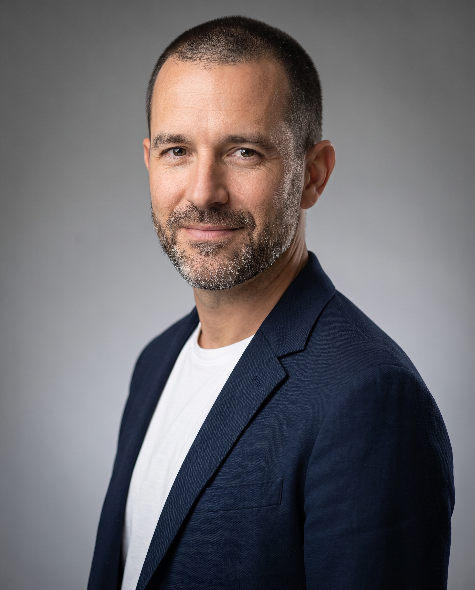 A corporate headshot of a bearded architect in a navy blazer on a soft grey studio background