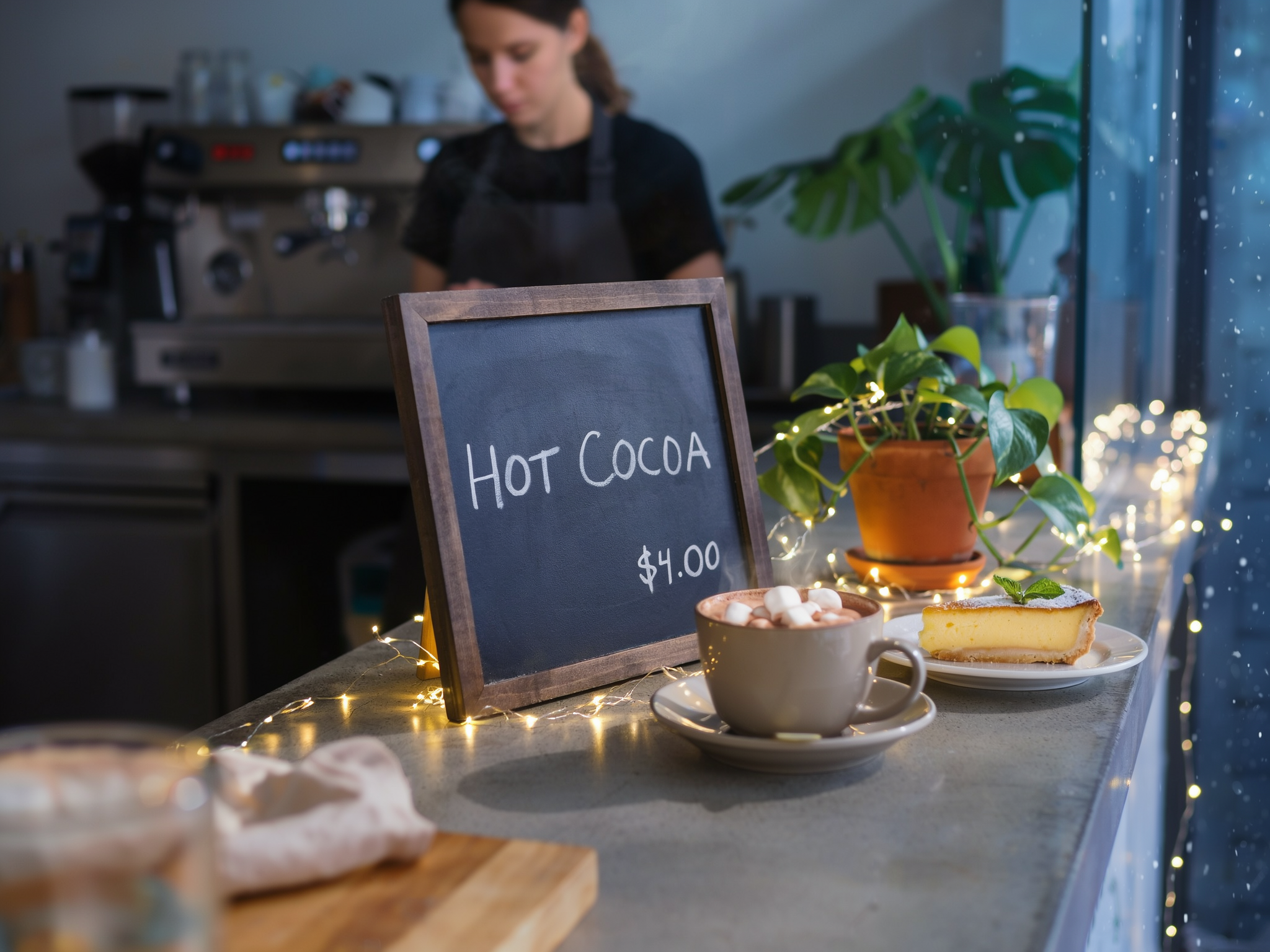 The same cafe counter turned into a winter evening: the chalkboard now reads HOT COCOA and $4.00, a mug of hot chocolate with marshmallows, fairy lights along the counter and snow falling outside the window