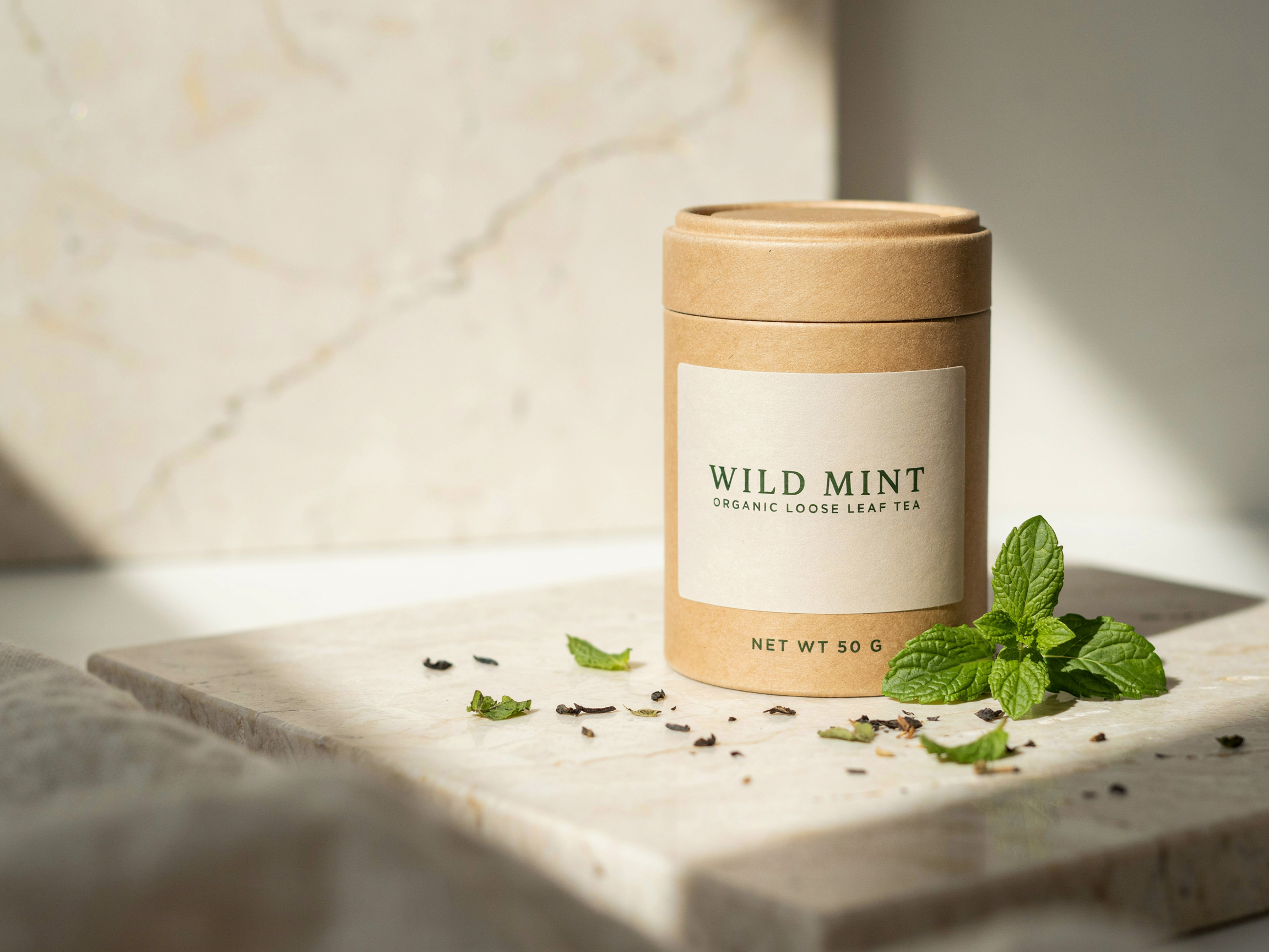 Kraft paper tea tin on pale travertine with a cream label reading WILD MINT, ORGANIC LOOSE LEAF TEA and NET WT 50 G, fresh mint sprigs and loose leaves around it in soft window light