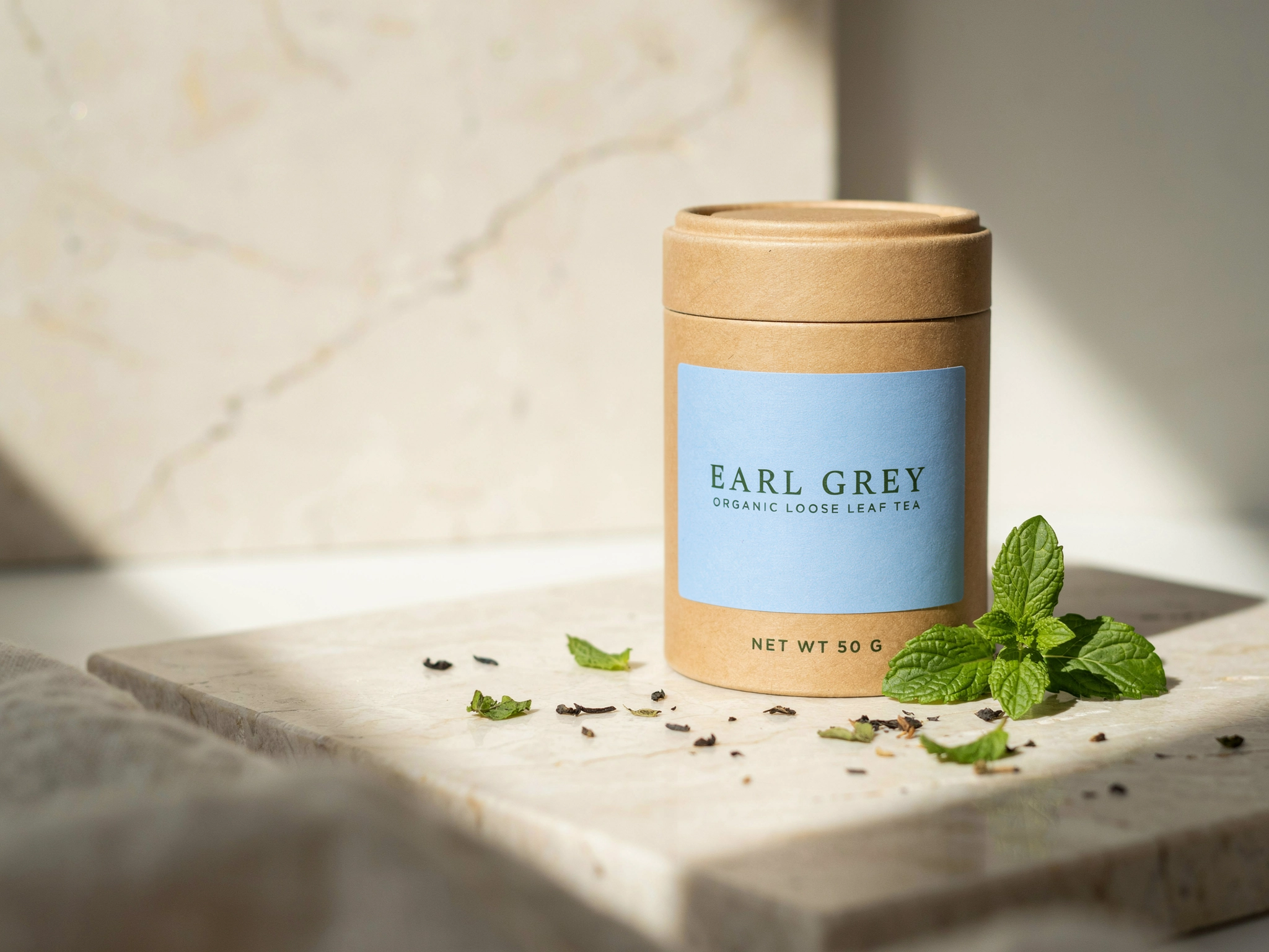 Precise Edit of the tea tin: the label is now pale blue and reads EARL GREY in green serif, with the same tin, travertine, mint and window light