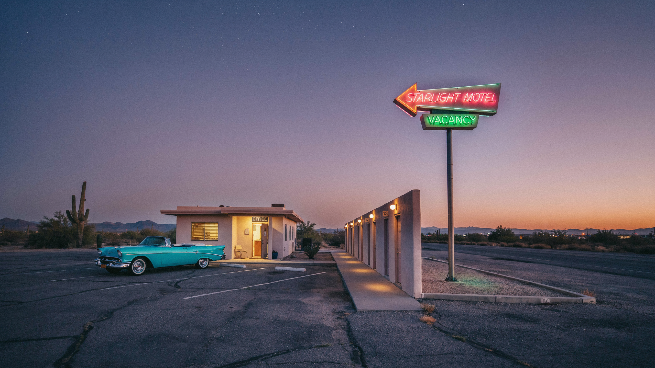 Dusk photo of an arrow shaped neon sign reading STARLIGHT MOTEL over a green VACANCY box, a turquoise convertible beside a small lit office, a row of lamplit doors and a saguaro cactus under a tangerine horizon