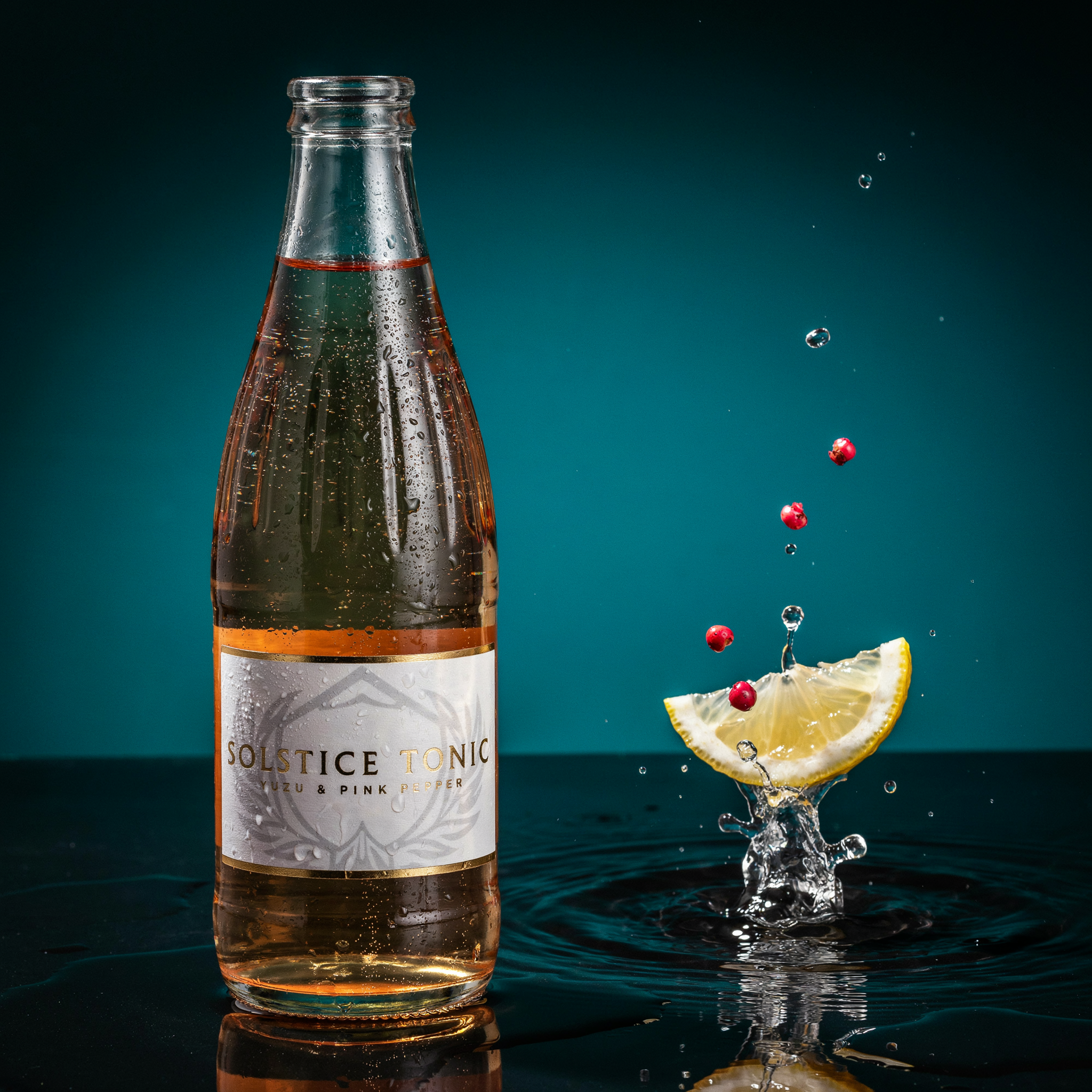 Glass bottle of pale amber tonic with a white label reading SOLSTICE TONIC and YUZU & PINK PEPPER in gold, beside a citrus wedge splashing into dark water with pink peppercorns falling against a teal background