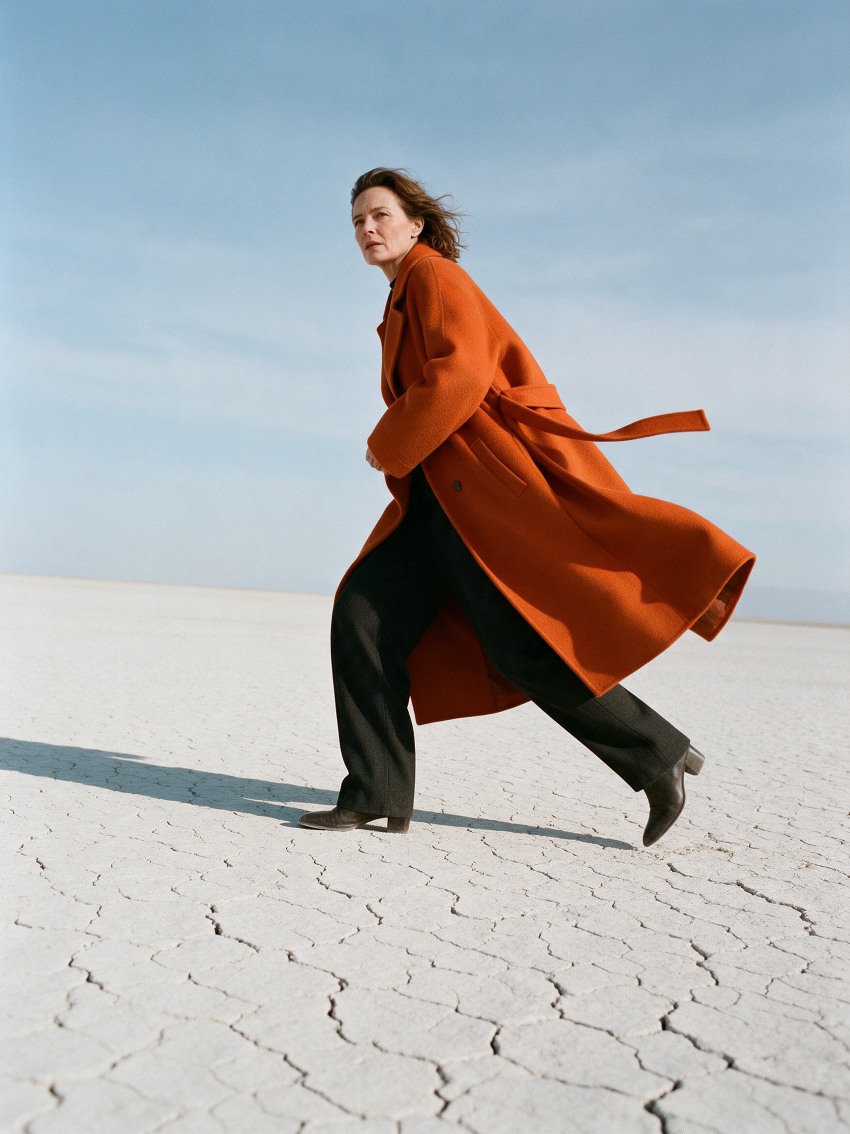 Seedream 5: the woman striding in profile across cracked white ground, the coat and its belt flying behind her