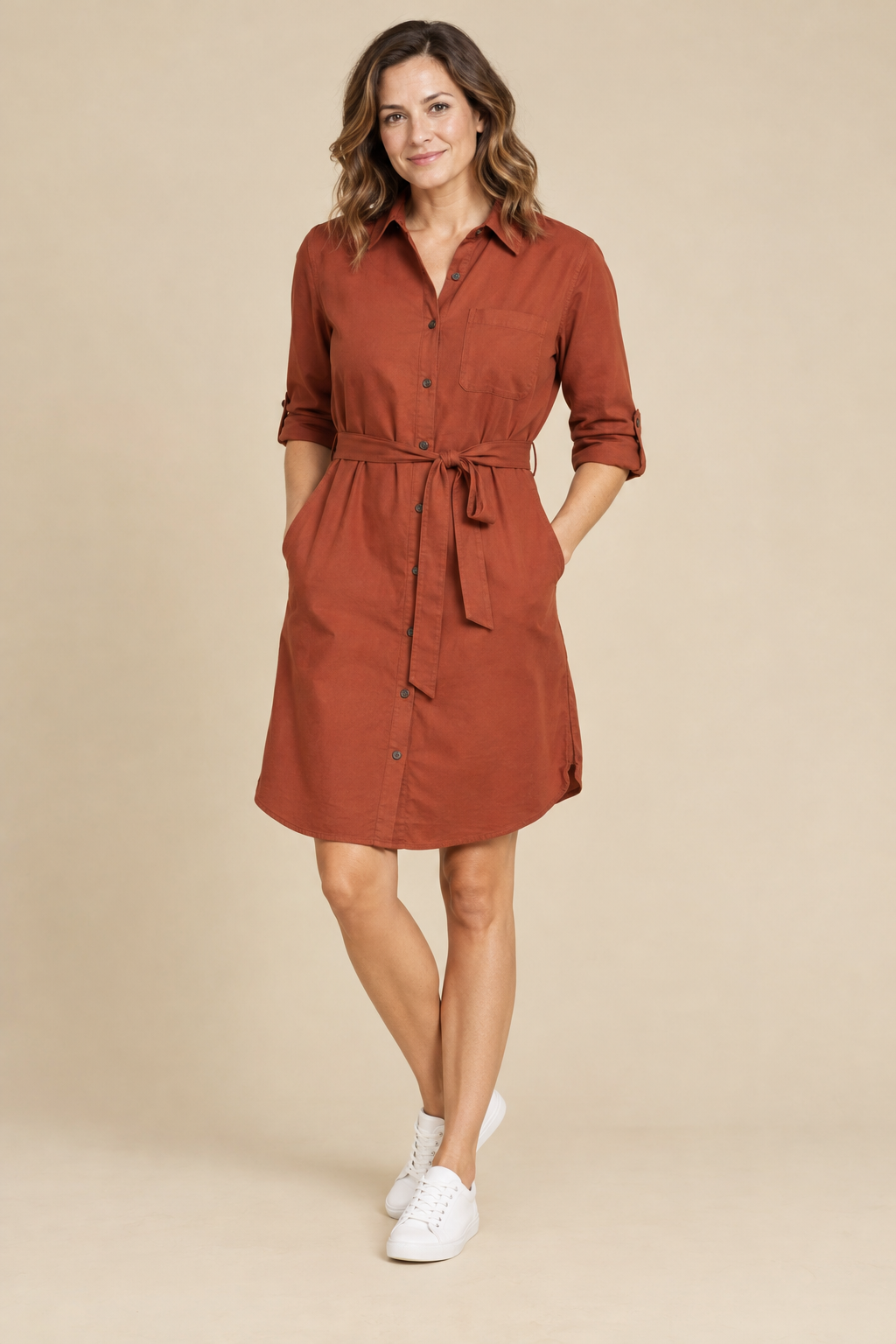 A woman in her thirties in a rust shirt dress and white sneakers stands against a sand colored studio backdrop