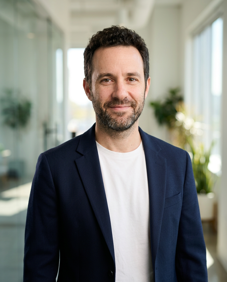 A man in his forties with curly dark hair and a beard in a navy blazer and white tee, smiling in a bright office with plants behind him