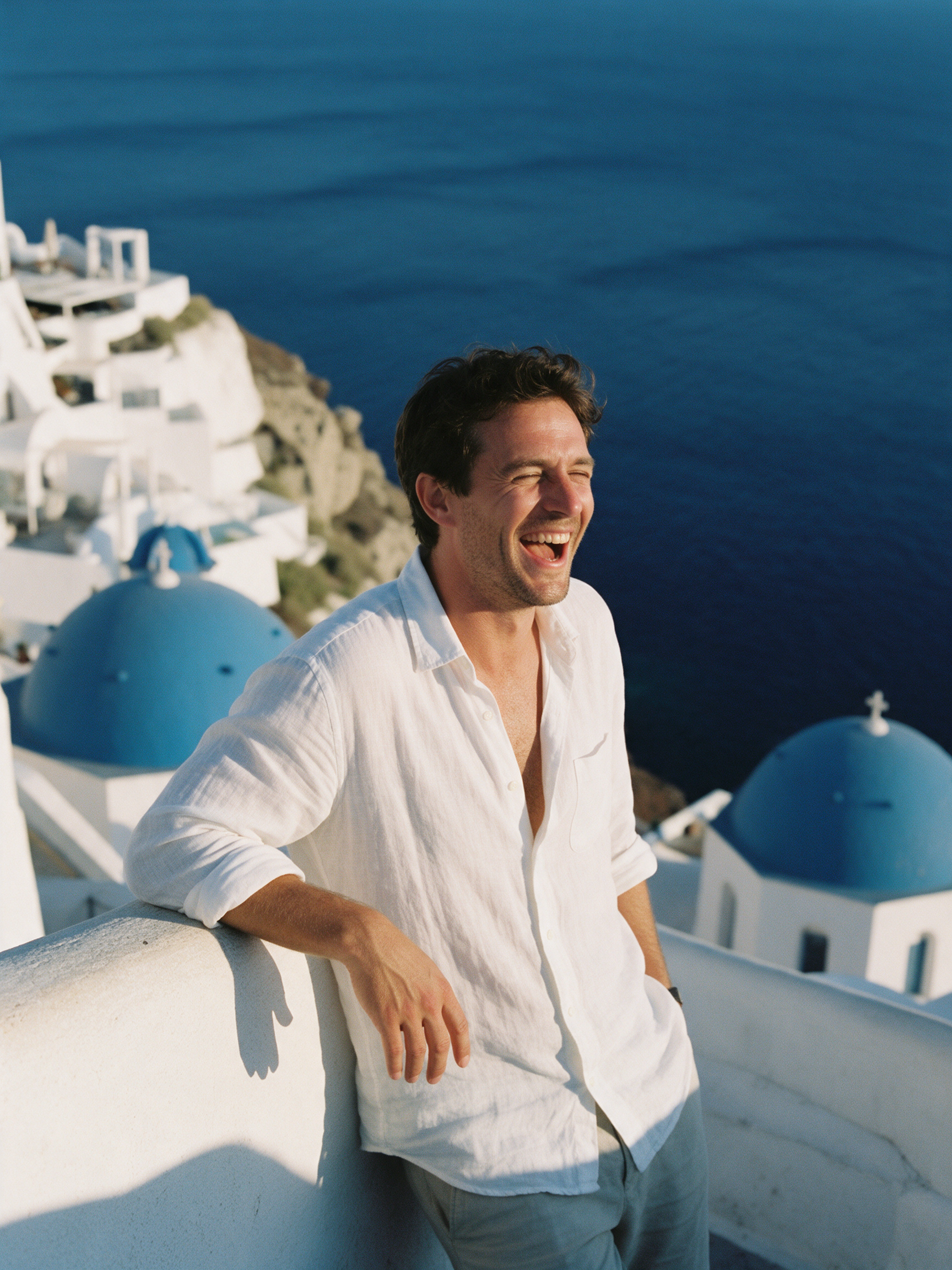 A laughing man in a white linen shirt leans on a whitewashed terrace wall above a deep blue sea with blue domed roofs below