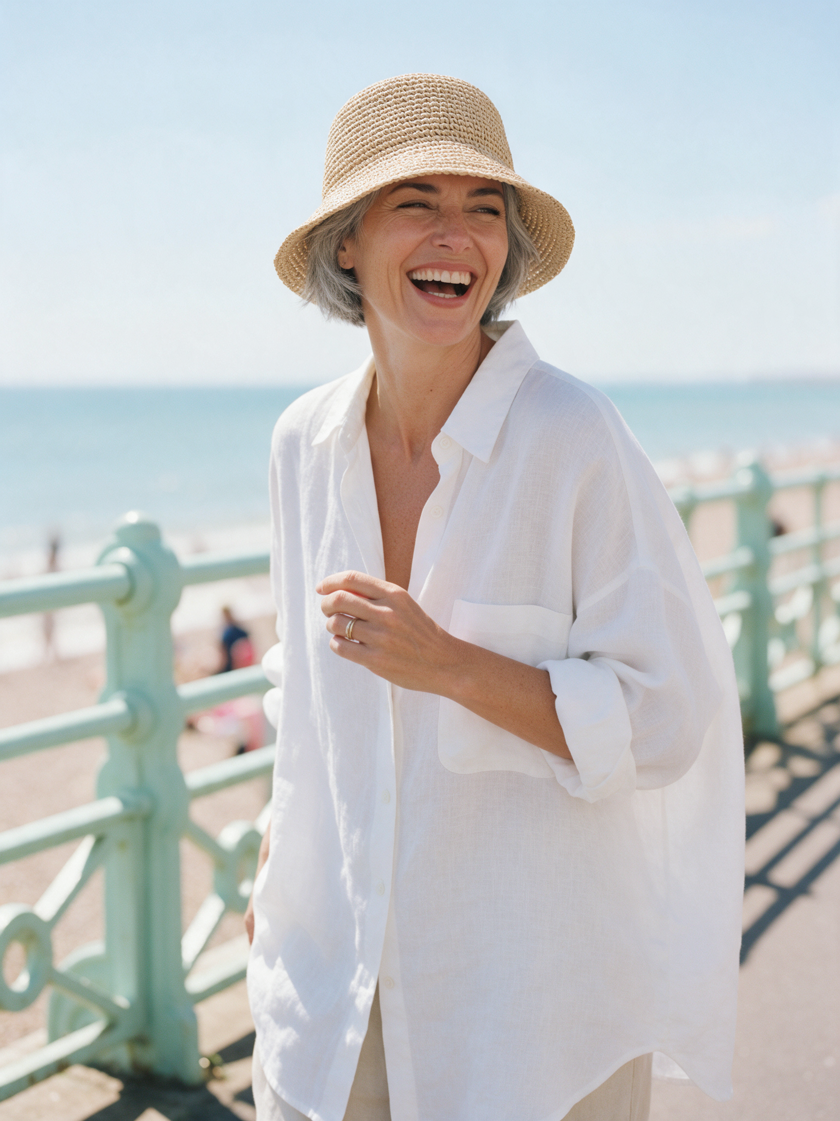 A laughing woman with grey cropped hair in a straw bucket hat and white linen shirt on a seaside promenade