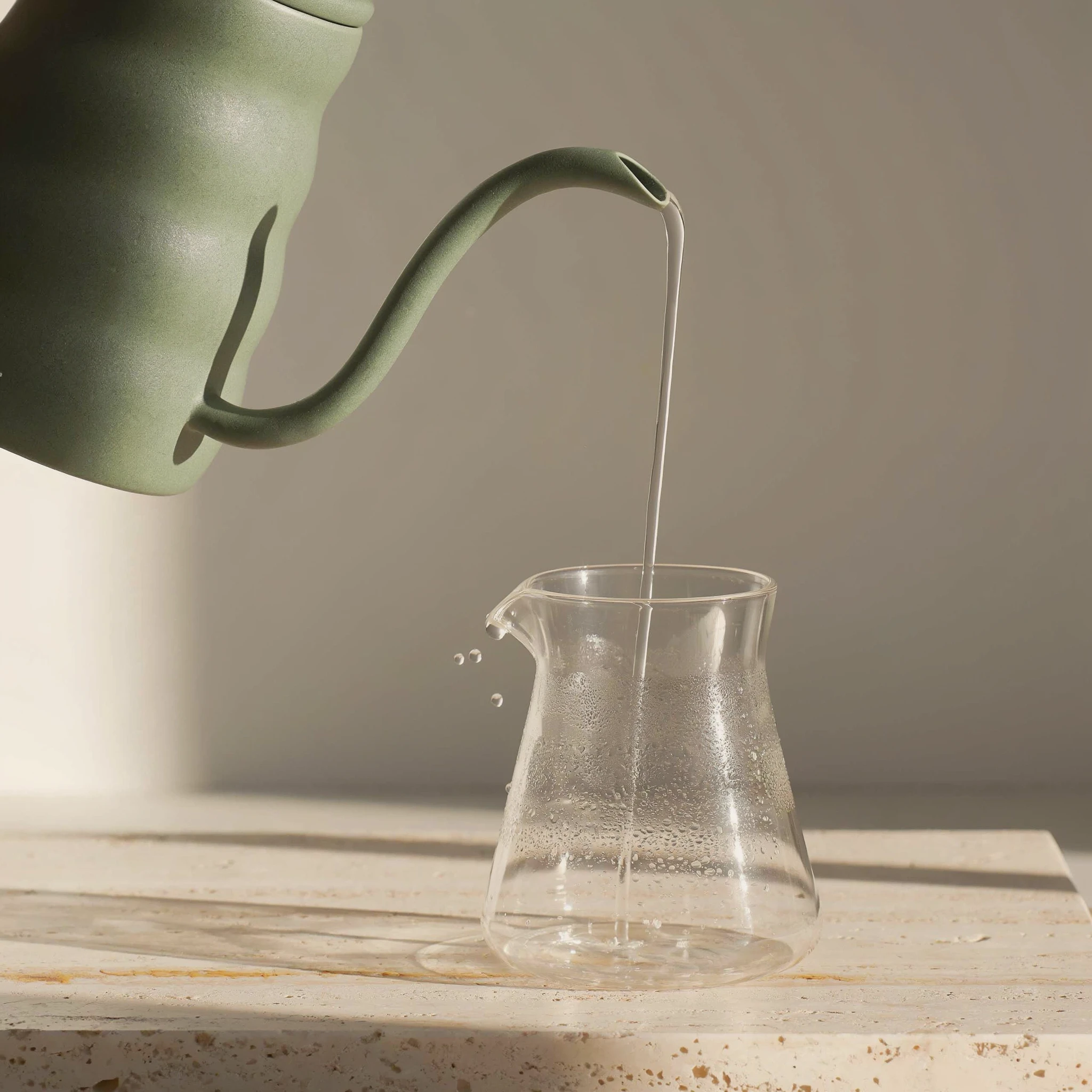 Sage green matte ceramic gooseneck kettle entering the frame from the top left and pouring a thin clear stream into a glass carafe beaded with condensation on a travertine slab, droplets flying from the carafe lip in raking morning light