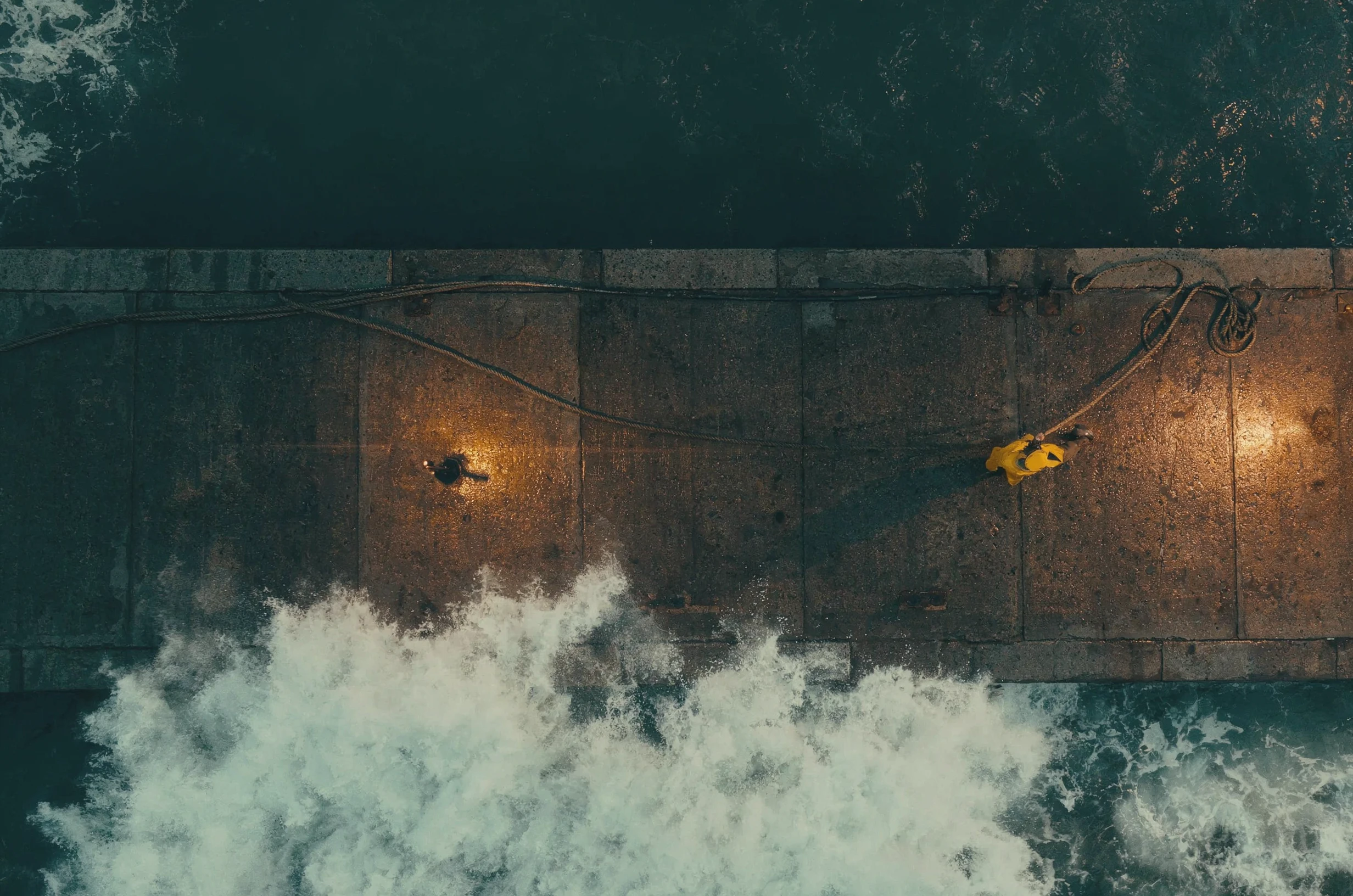 Top down drone view of a wet concrete pier at dusk: a small figure in a yellow coat hauls a rope coiled at the pier edge, warm pools of lamplight on the stone and a white wave breaking along the bottom of the frame