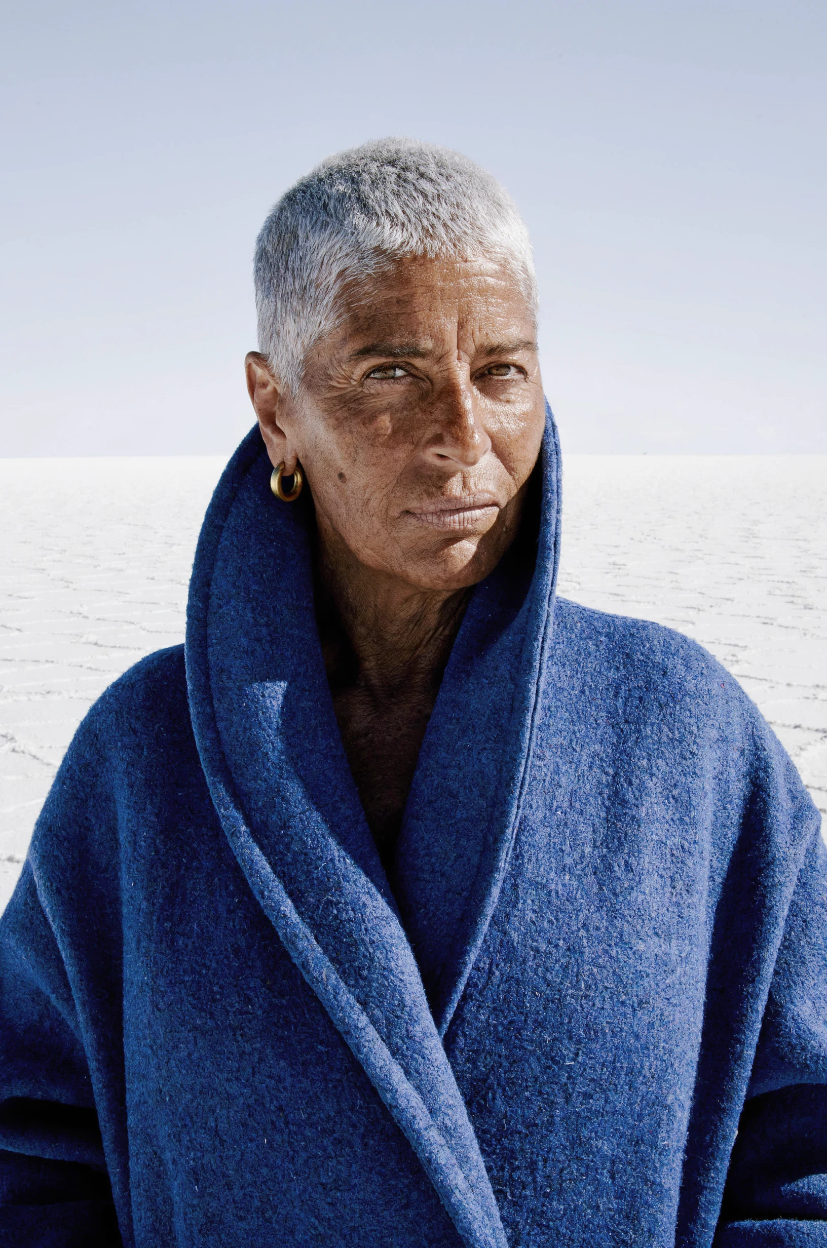 Luma UNI 1 MAX portrait: a woman in her sixties with a silver buzz cut, deeply lined sun spotted tan skin and a gold hoop earring, wrapped in a high collared cobalt wool coat, facing the camera on a salt flat in hard light