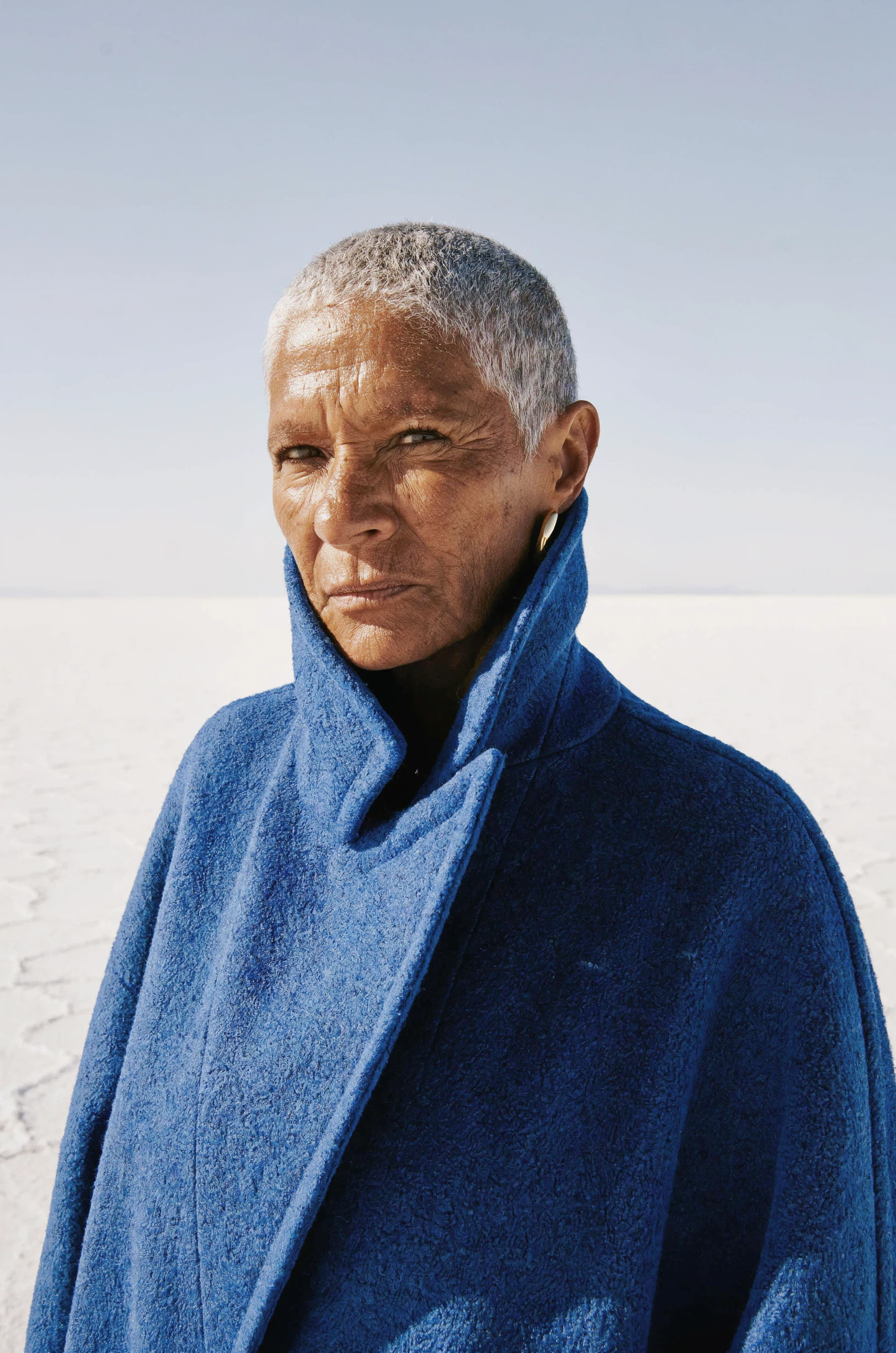 Close portrait of a woman in her sixties with a silver buzz cut and deeply lined tan skin, a gold earring, the high collar of a cobalt blue wool coat turned up around her face, a white salt flat and pale sky behind her in hard sun