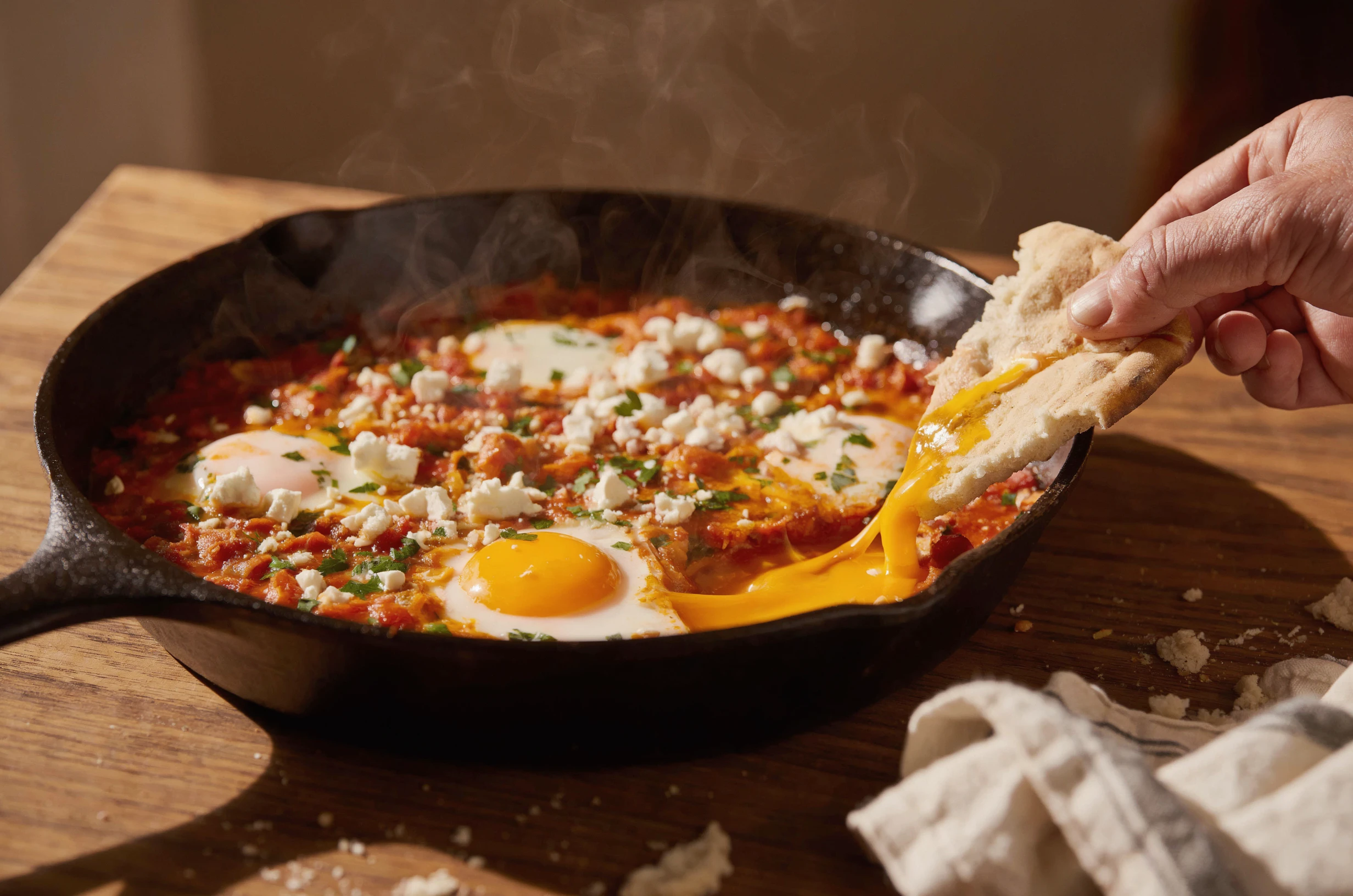 Cast iron pan of shakshuka with crumbled feta and parsley on a walnut table, a hand dragging torn flatbread through a runny yolk that streams in a long golden drip, steam rising in warm side light, a linen napkin in the corner