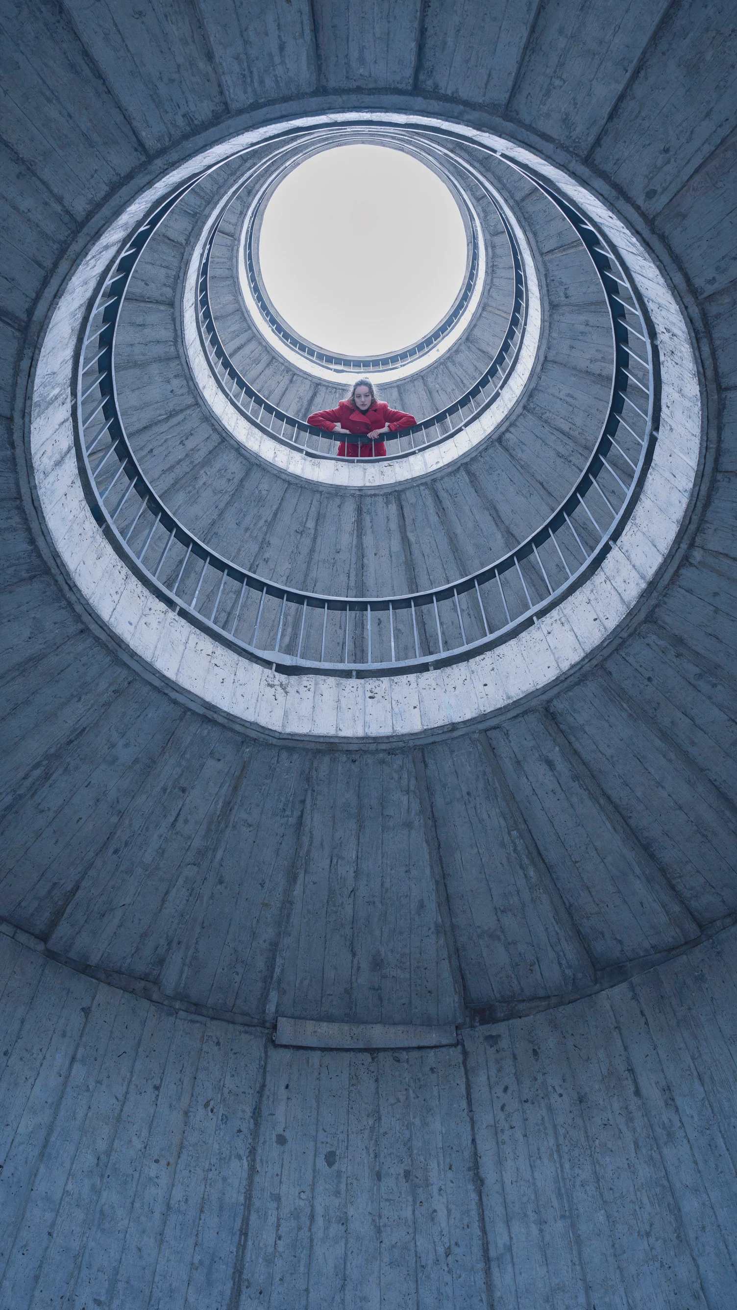 Looking up a ringed concrete shaft toward a round white skylight, stacked railings circling above, a woman in a red wool coat leaning on a railing with folded arms and looking down at the camera, cool grey concrete filling the frame