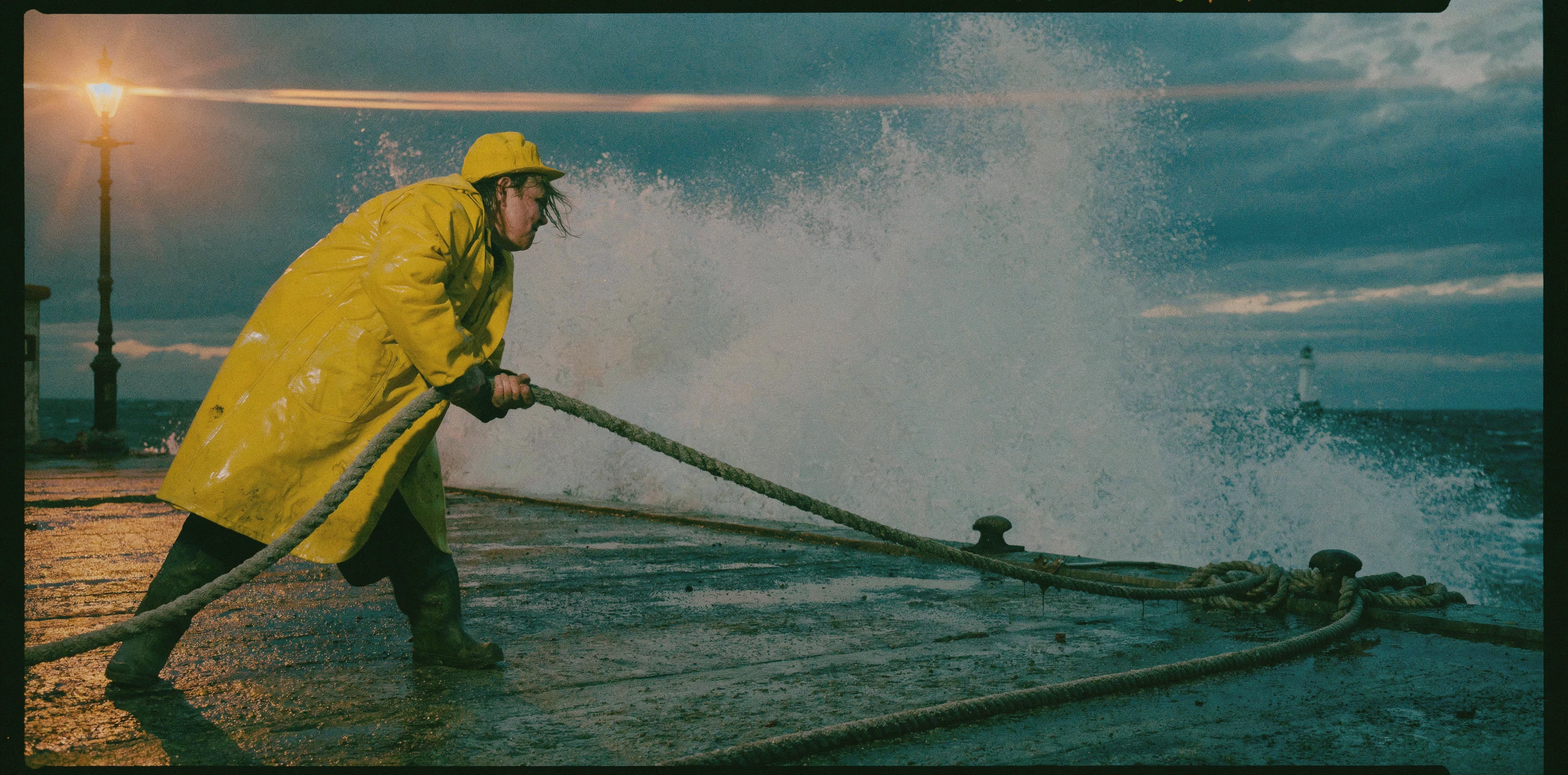 Wide film still with a black frame border of a woman in a yellow oilskin coat and cap leaning forward to pull a rope on a wet pier, a wave exploding into spray behind her, a lit street lamp at left and a small lighthouse on the horizon