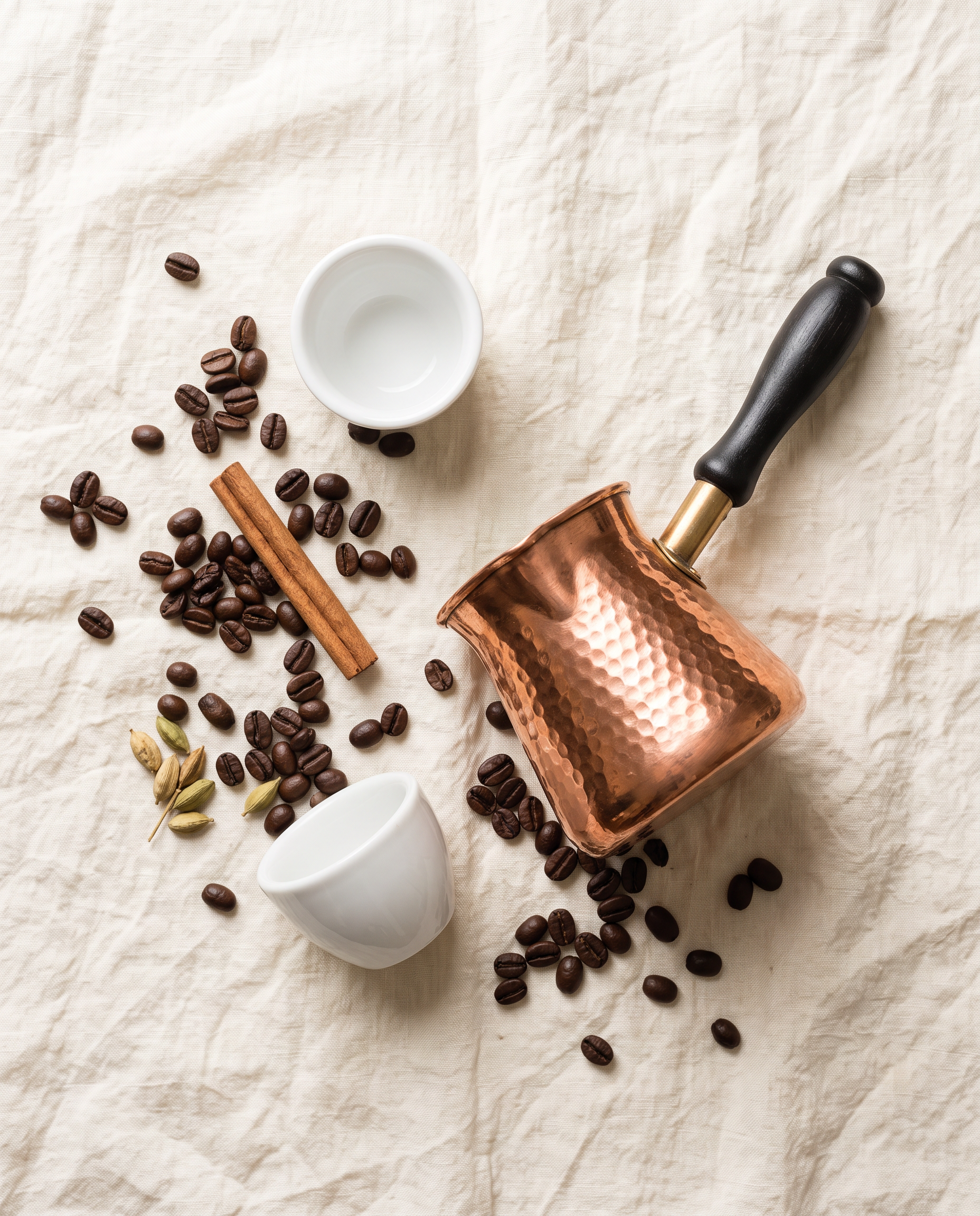 An overhead flat lay of the copper coffee pot on linen with cups, beans and spices