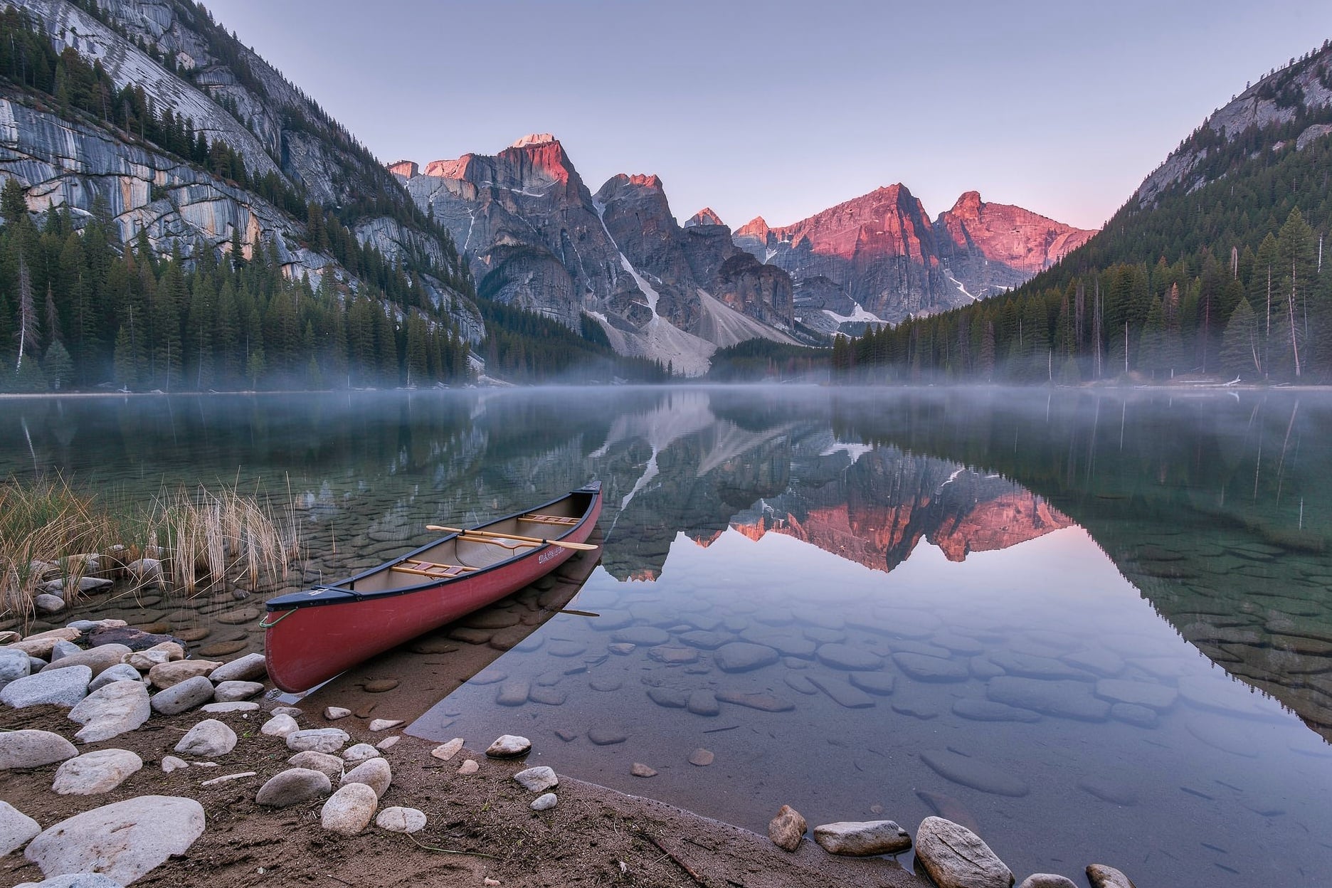 A red canoe on a still alpine lake at dawn with peaks mirrored in the water