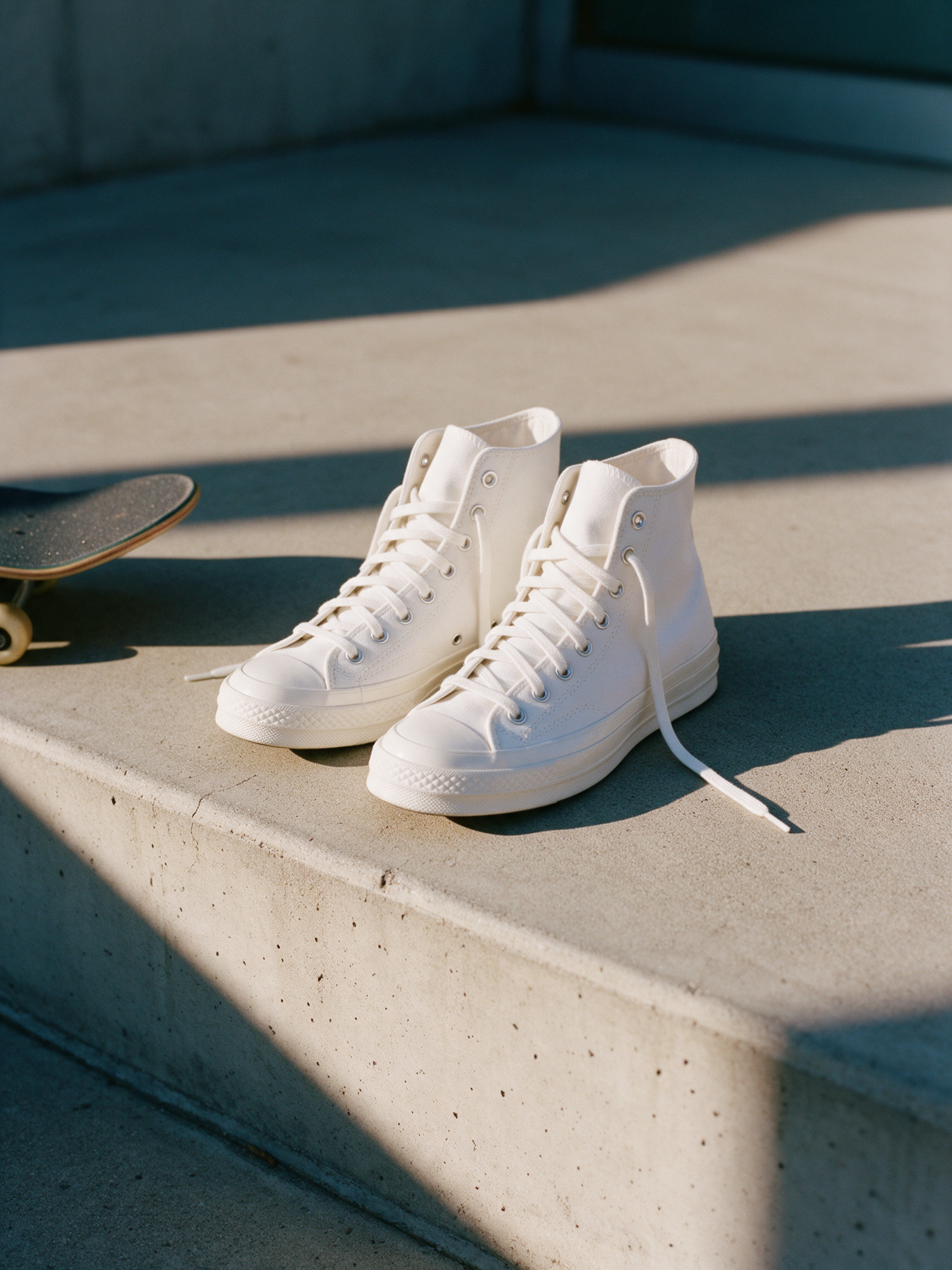 White canvas high top sneakers stand on a sunlit concrete step beside the tail of a skateboard