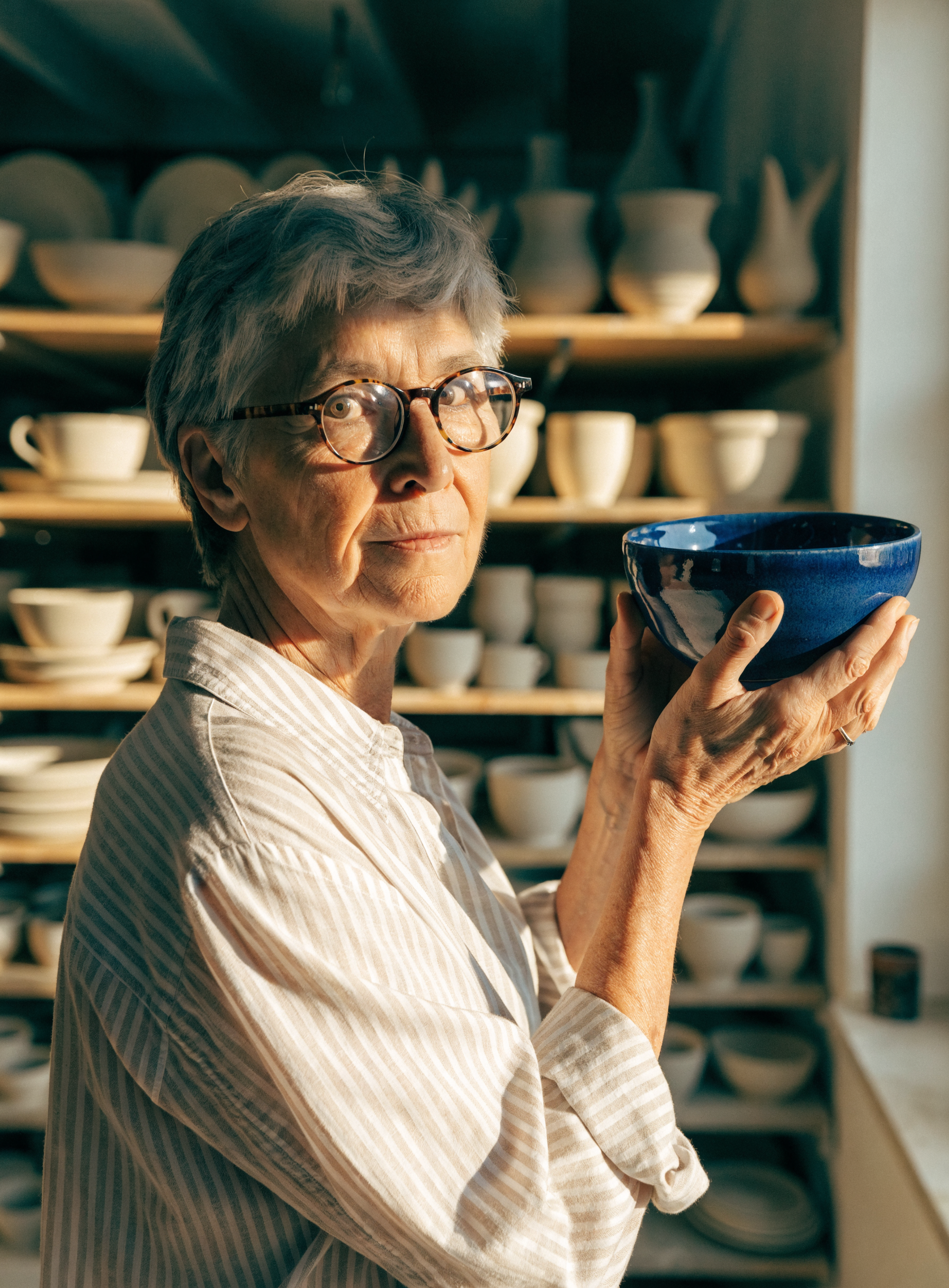 Photo of a woman in her sixties with short grey hair and round tortoiseshell glasses holding up a glossy cobalt blue bowl in a sunlit pottery studio lined with unglazed pots