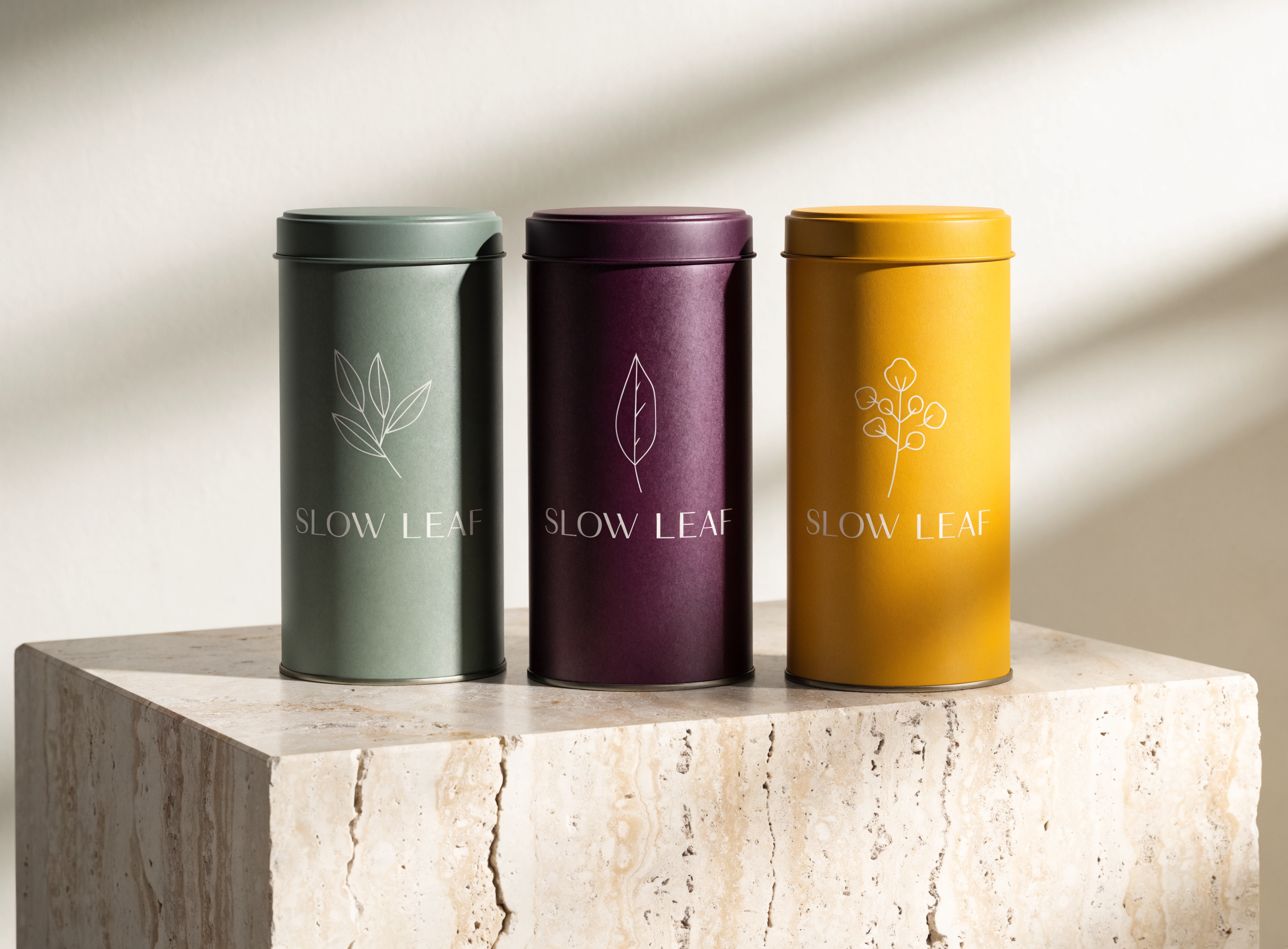 Three matte tea tins in sage green, plum and saffron yellow, each printed with a white botanical line drawing and the serif wordmark SLOW LEAF, standing on a travertine block in soft window light